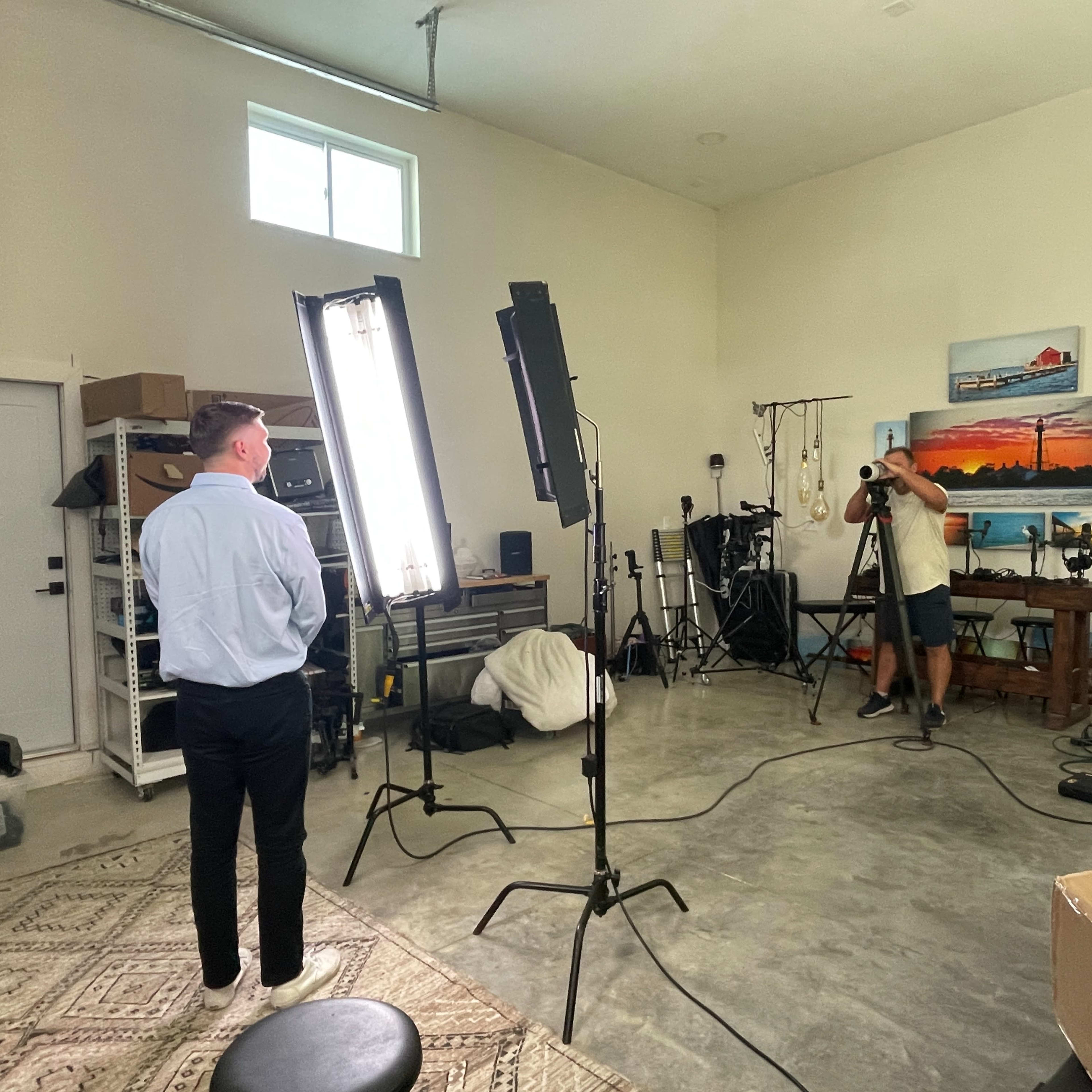 A person in a light blue shirt stands in front of two bright lights while another person holds a camera in a studio-like setting.