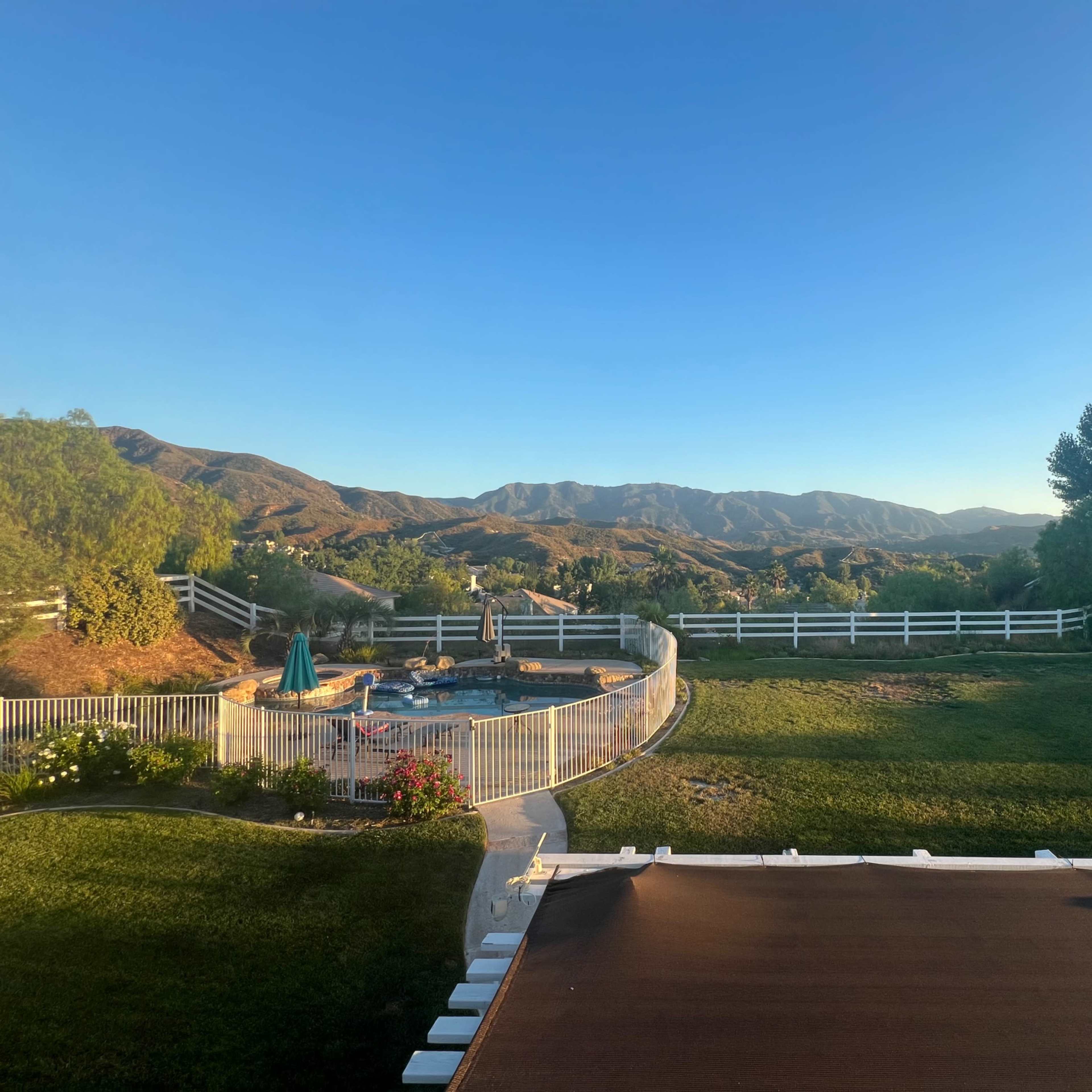 A backyard with a swimming pool surrounded by a fence, set against a backdrop of rolling hills and clear blue skies.