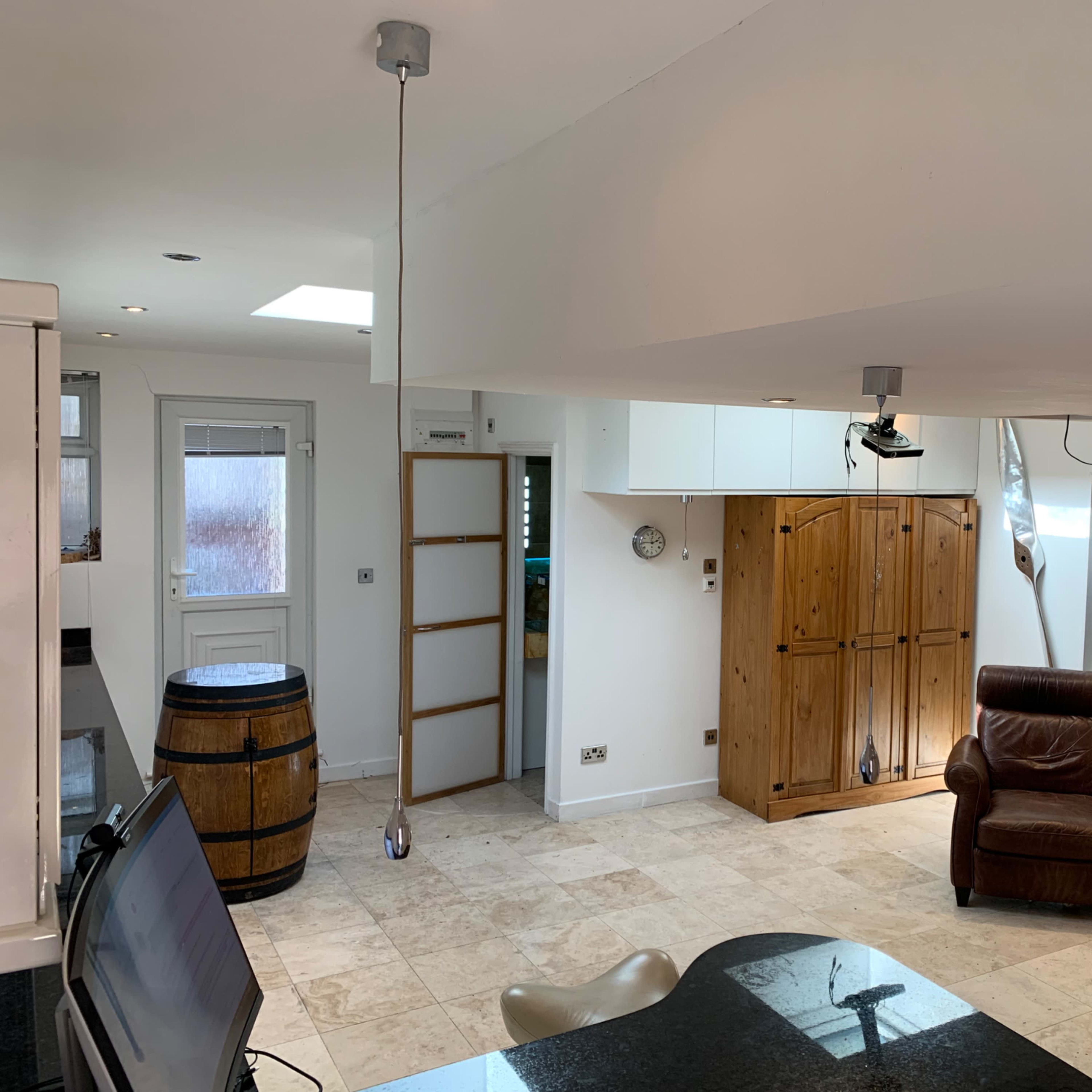 A modern, open-plan kitchen and living area with a leather armchair, a wooden cabinet, and a barrel, featuring bright white walls and tiled flooring.