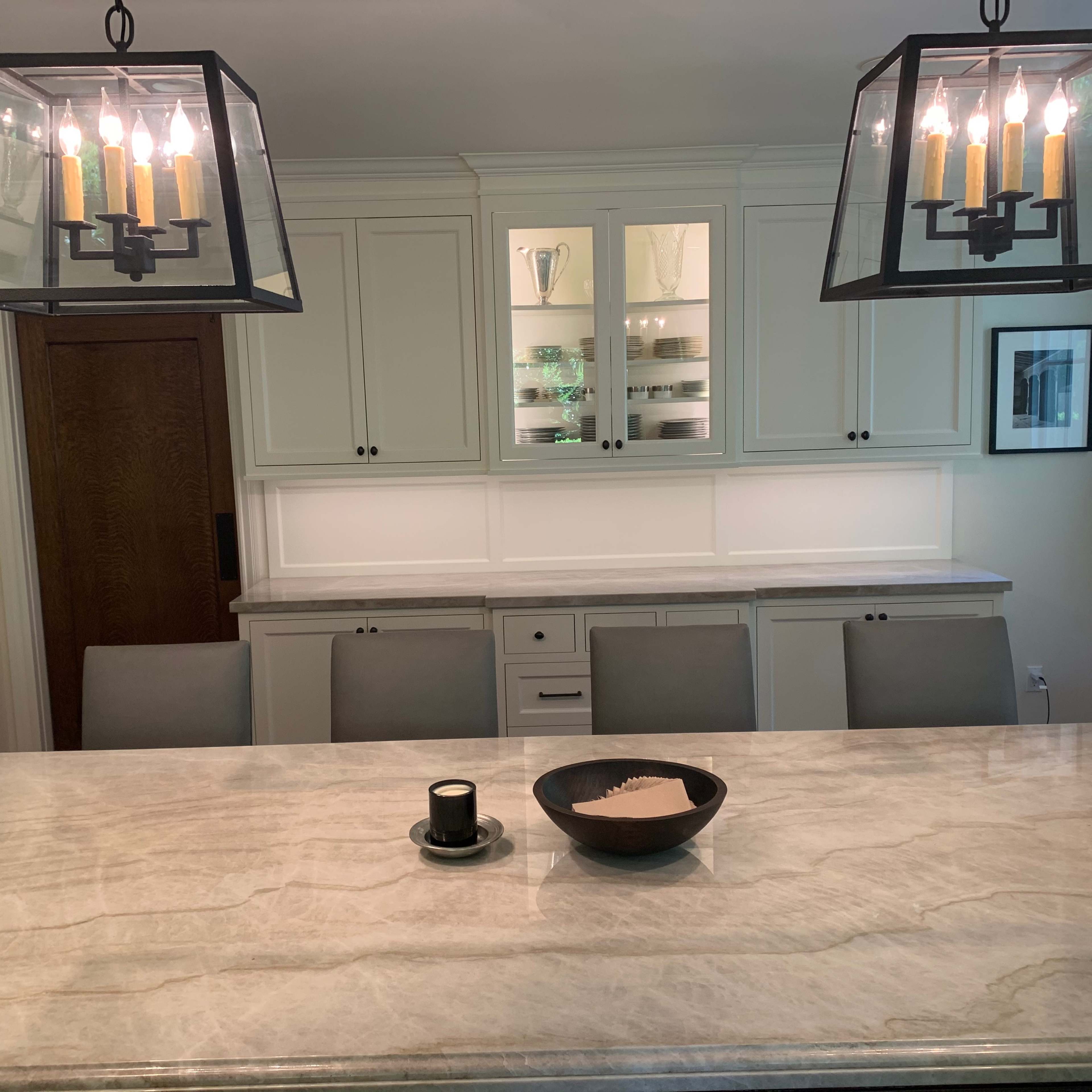 A modern kitchen features a large marble island, contemporary light fixtures, and a wall of white cabinetry with glass panels.