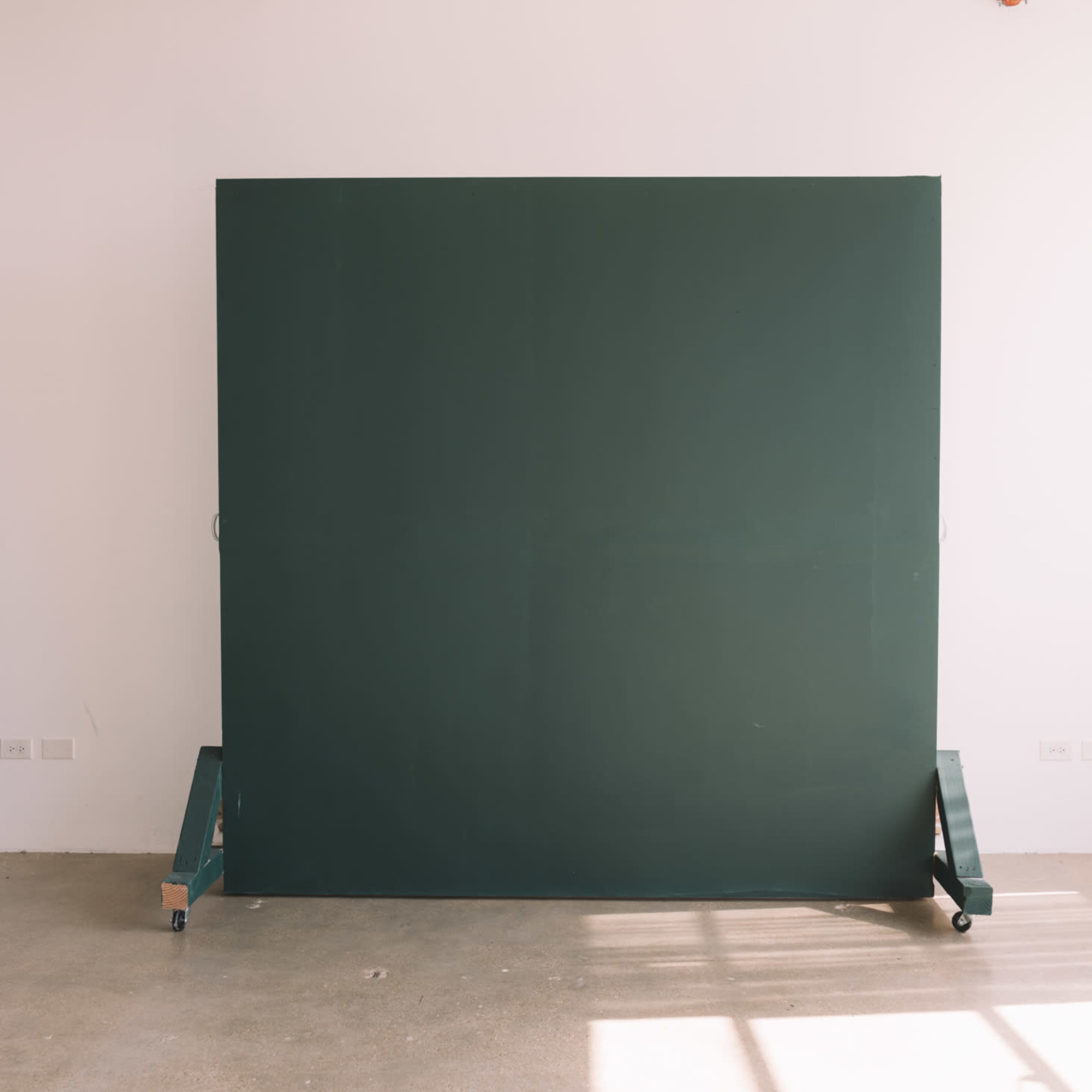 A large green board on wheels is positioned against a plain white wall in a spacious room with concrete flooring.