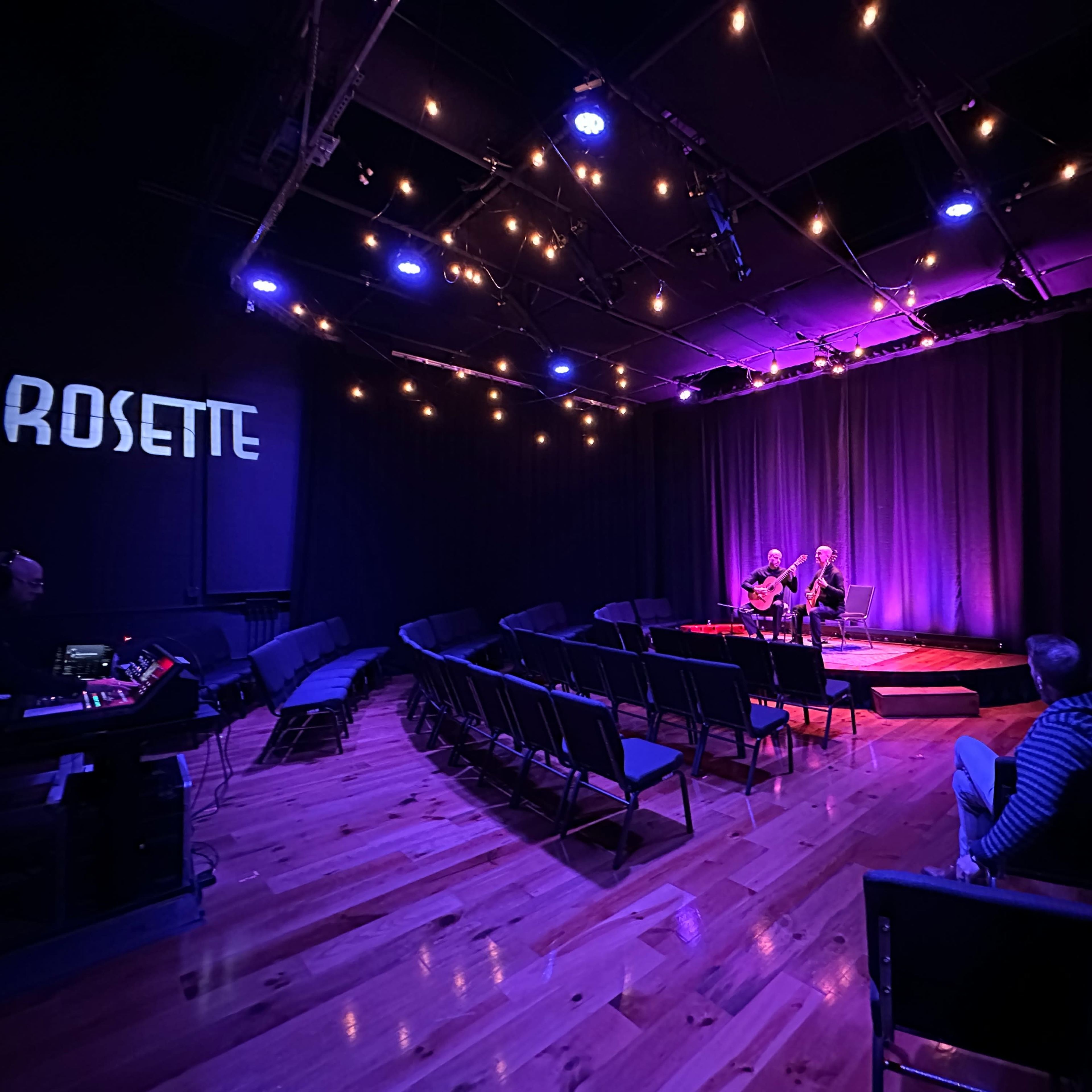 A dimly lit performance space with empty chairs facing a small stage where two musicians play, illuminated by purple and blue lights.