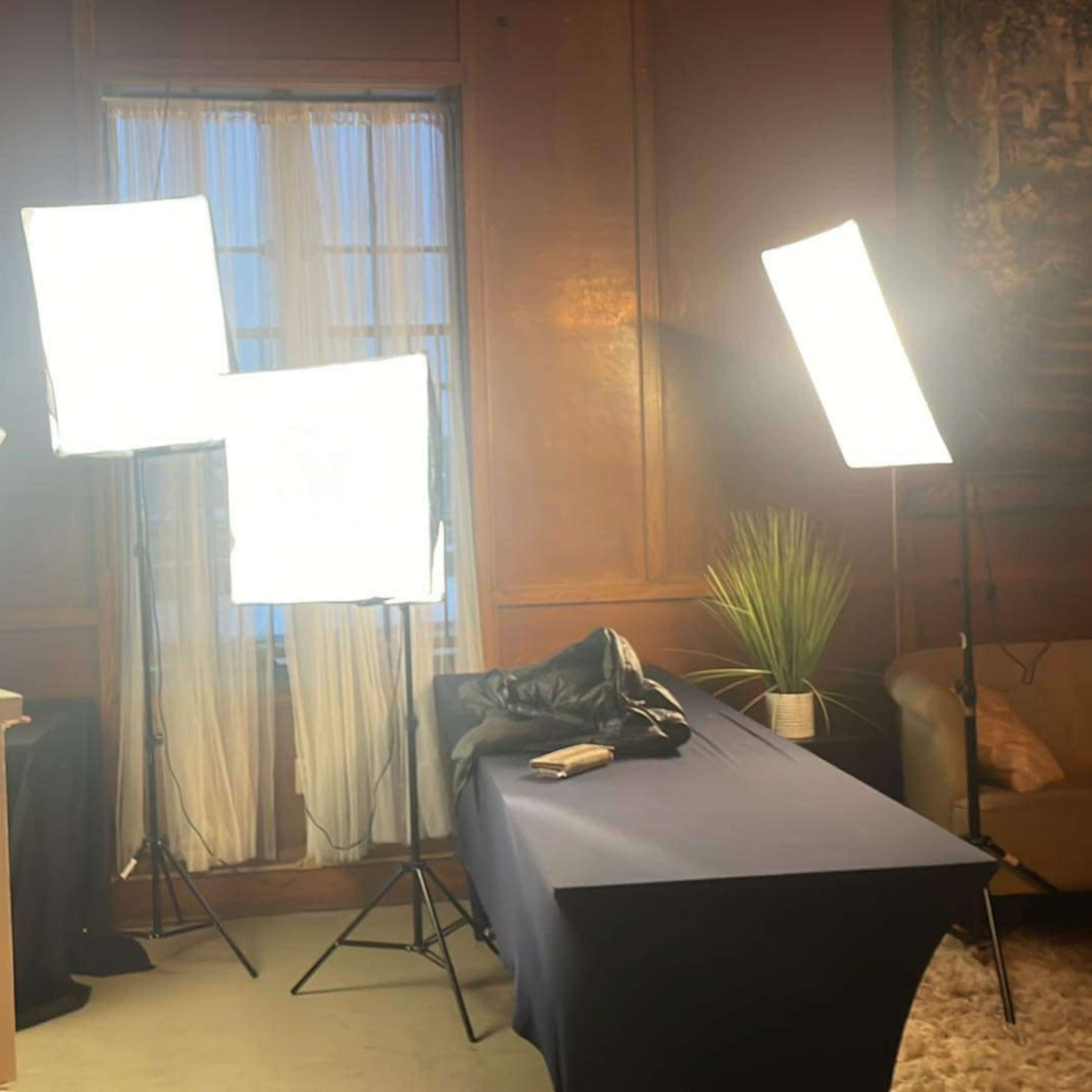 The scene features multiple softbox lights arranged around a table draped with a blue cloth in a room with a large window and a plant.
