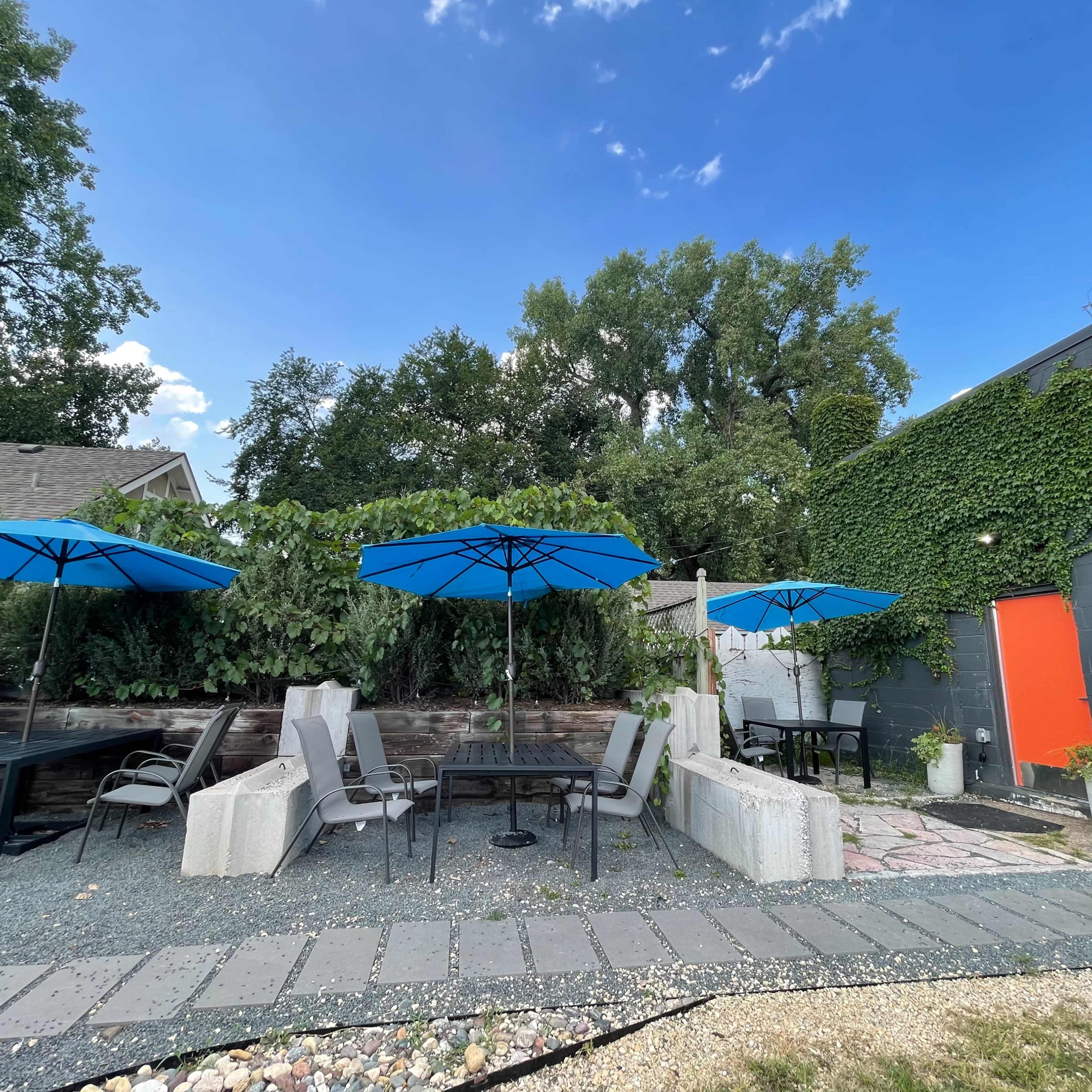 An outdoor patio area with several tables and chairs under blue umbrellas, surrounded by greenery and a colorful wall.