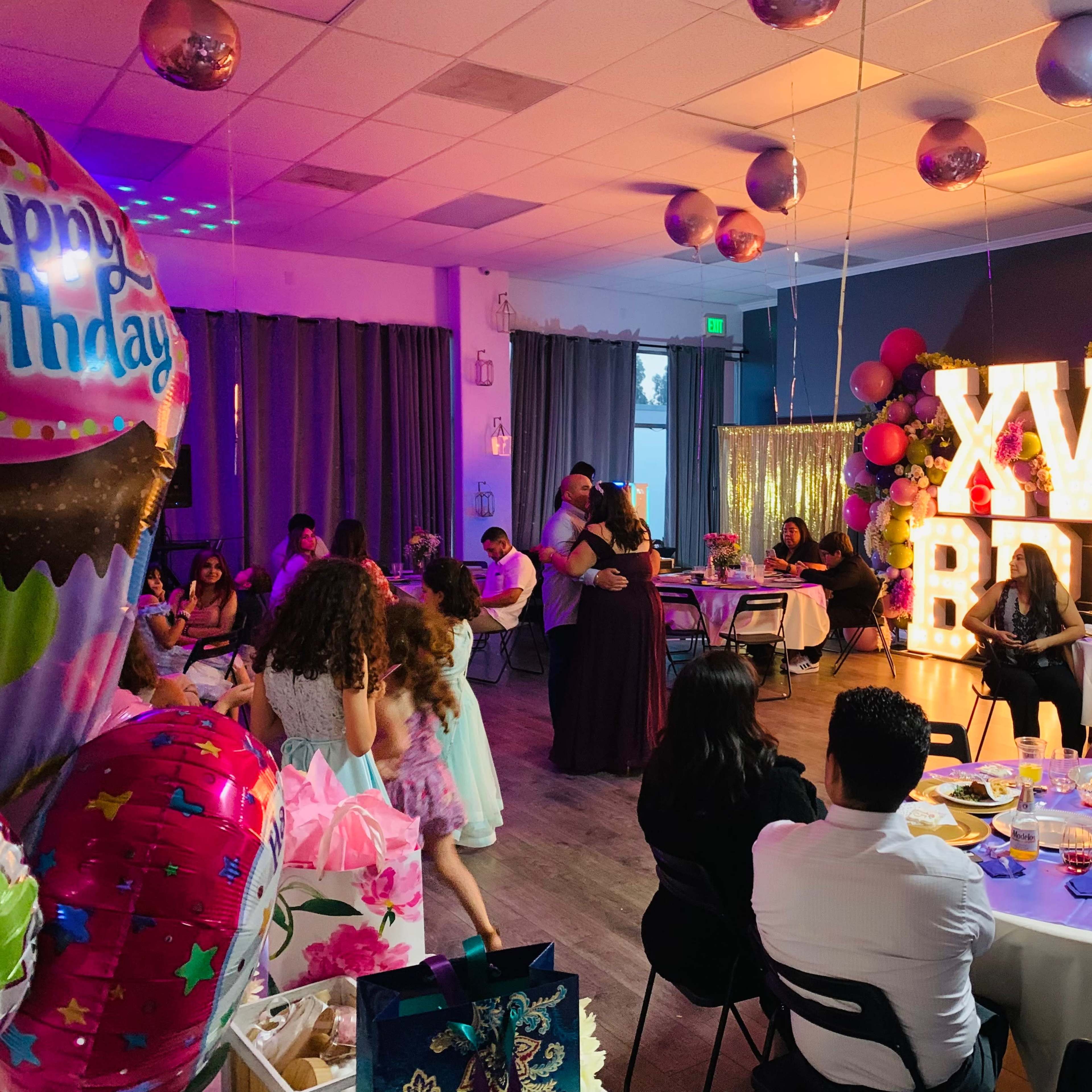 A celebration scene featuring a large "XV BD" marquee sign, with guests enjoying food and dancing at a birthday party.