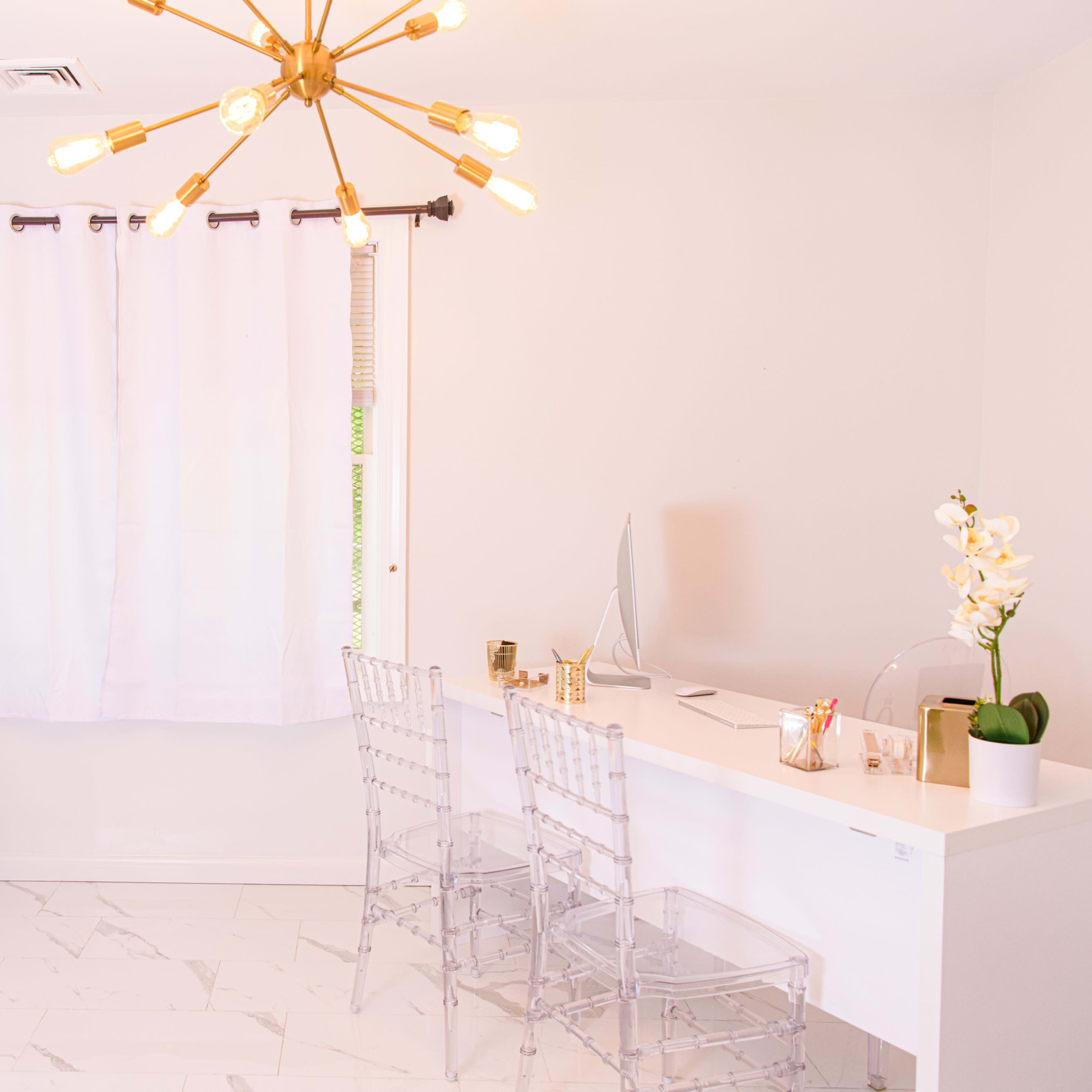 A modern workspace features a white desk with two clear chairs, a large mirror, and a pendant light, all set against a bright, minimalist backdrop.
