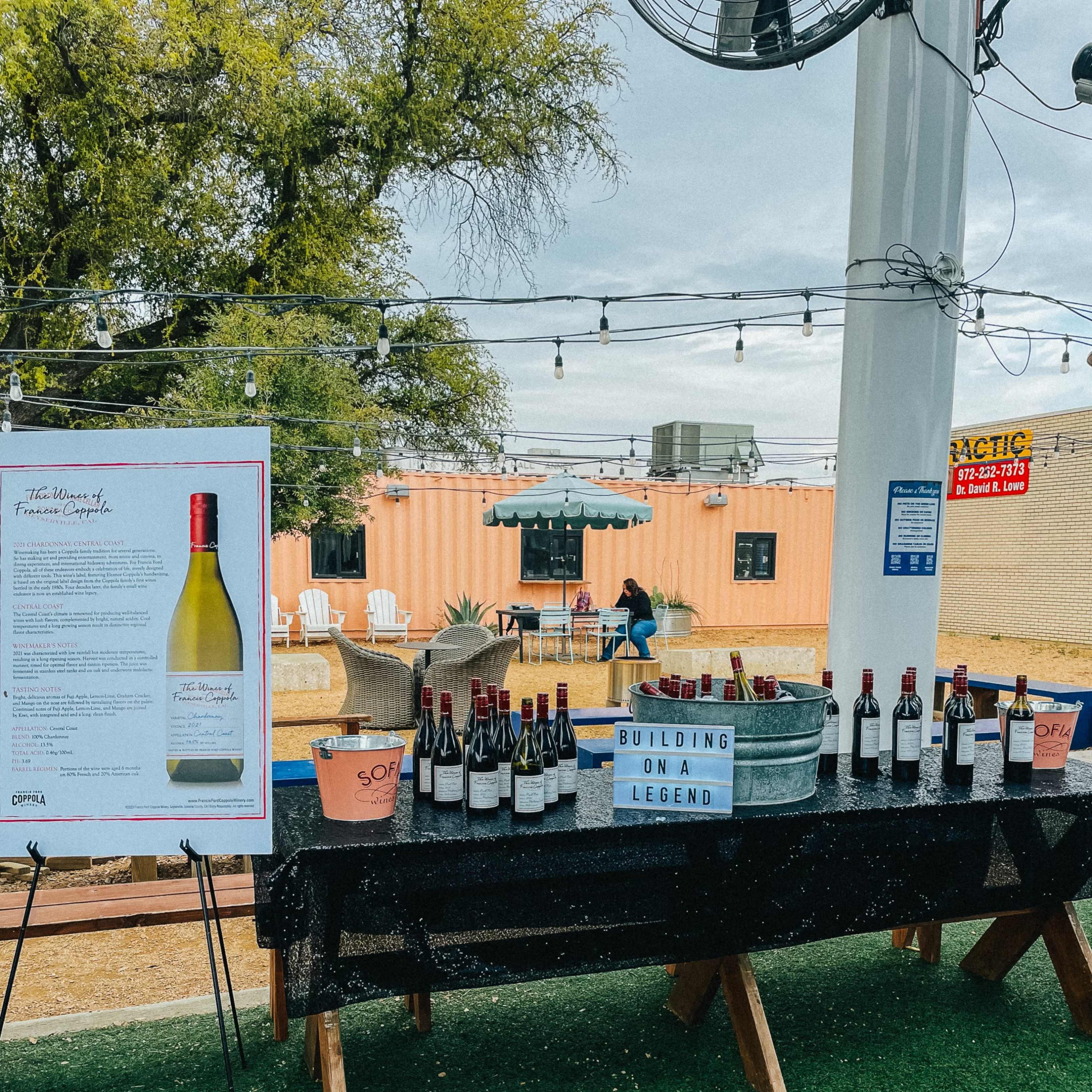 A display table featuring multiple bottles of wine, with a sign and a container of ice, is set up in a outdoor venue with string lights overhead.