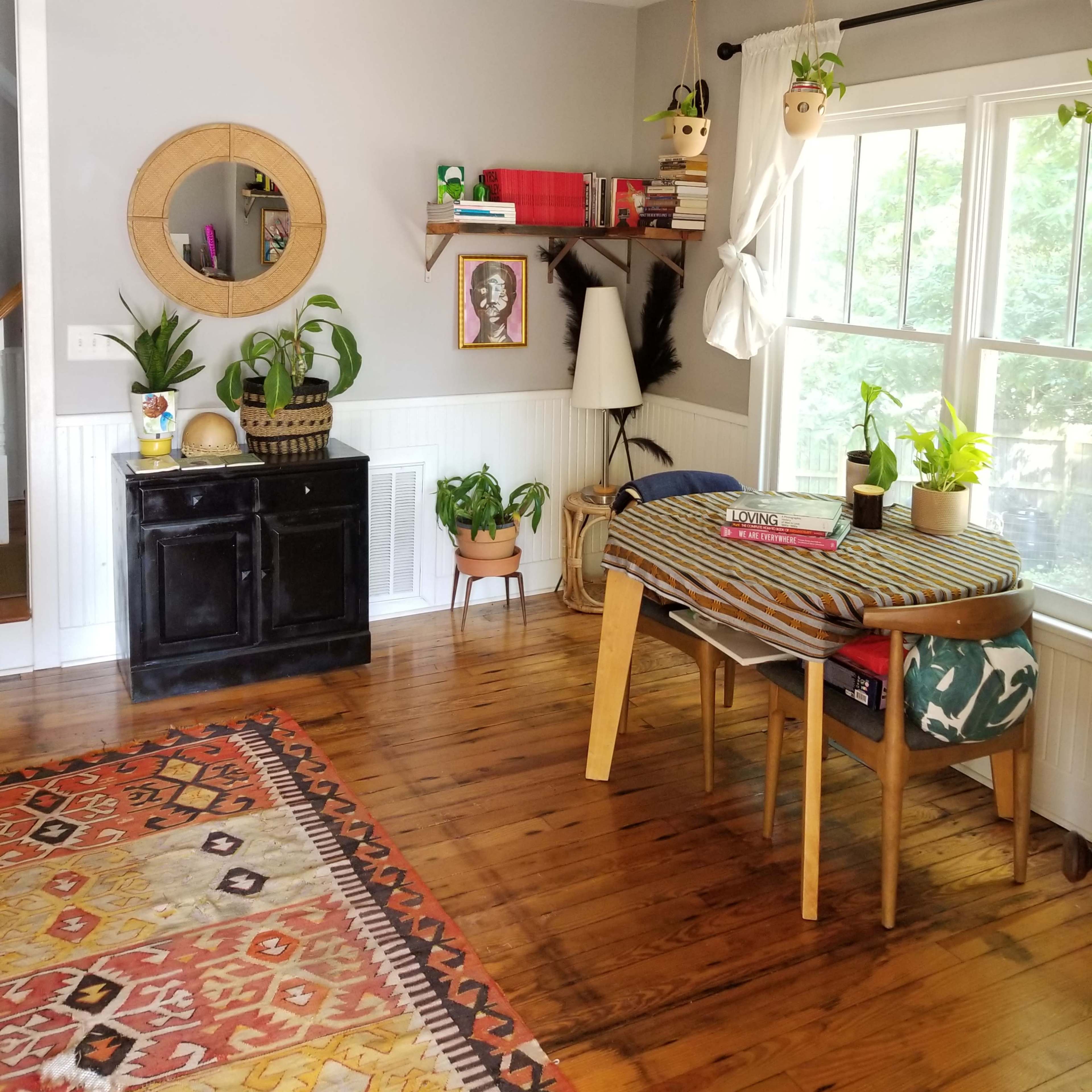 The scene features a cozy room with wooden flooring, a black cabinet, a window with plants, and a small table stacked with colorful items.