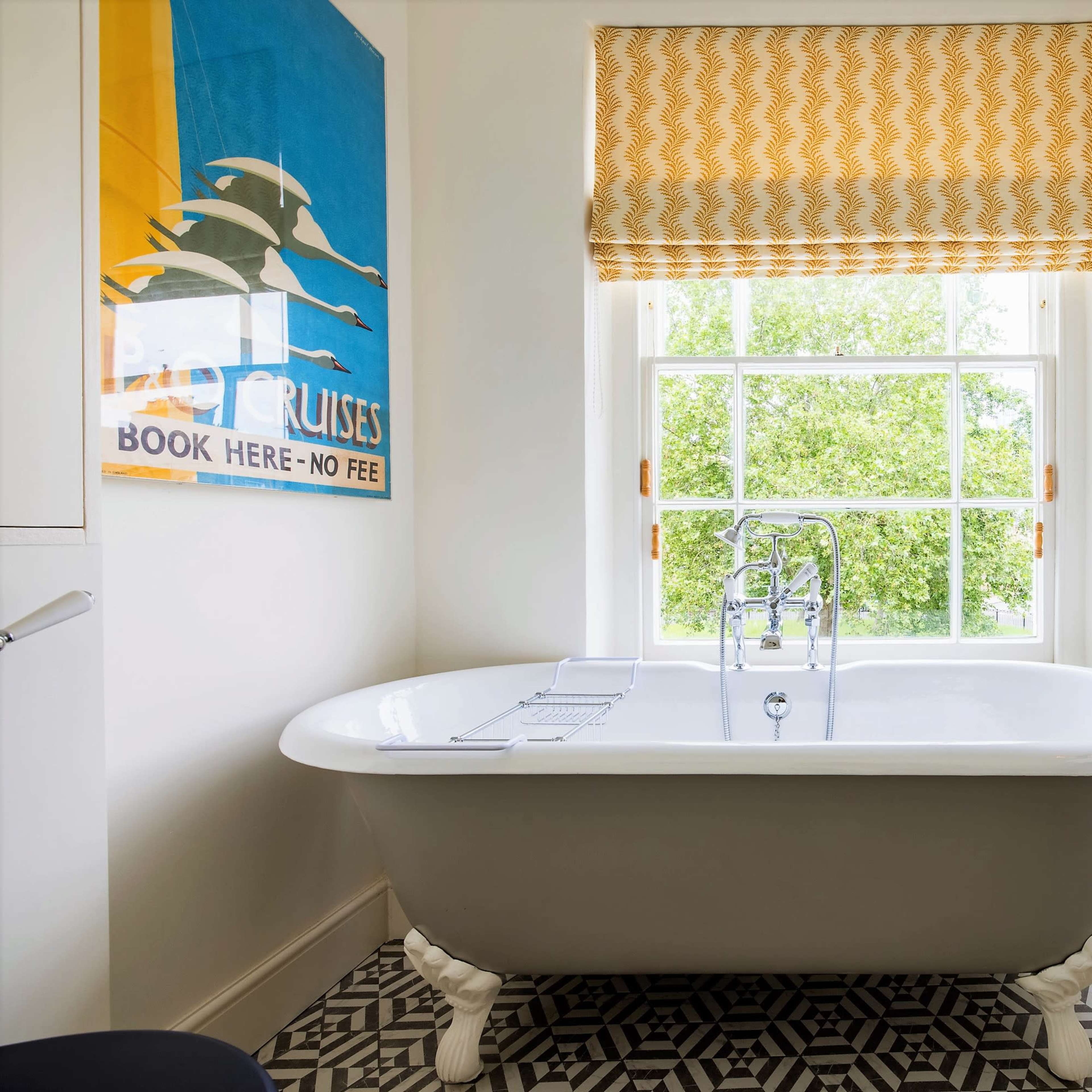 A bright bathroom featuring a freestanding white bathtub under a window with a yellow patterned shade, alongside a sink and decorative wall mirror.