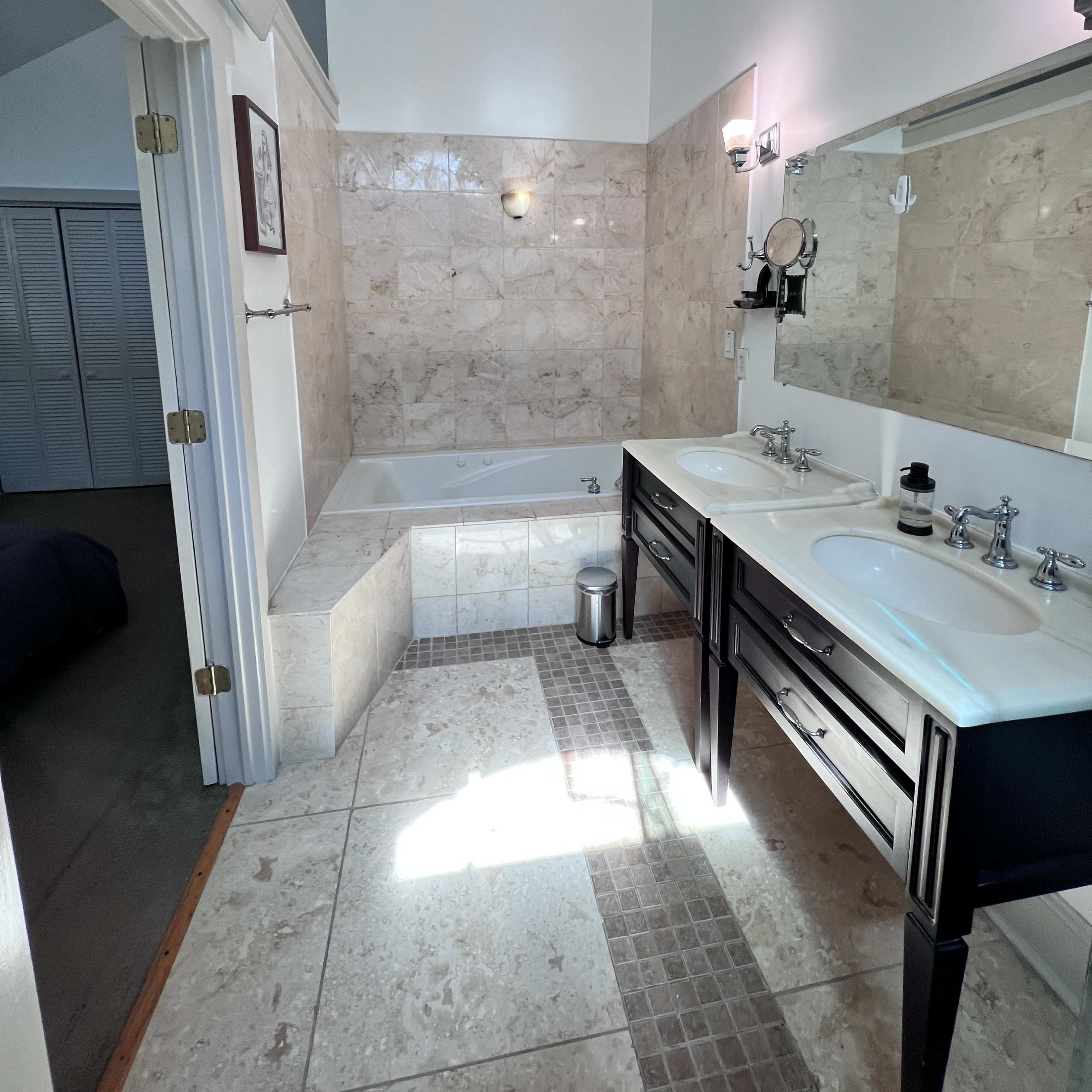 A bathroom featuring a double vanity with sinks, a bathtub, and a tiled floor, illuminated by natural light.
