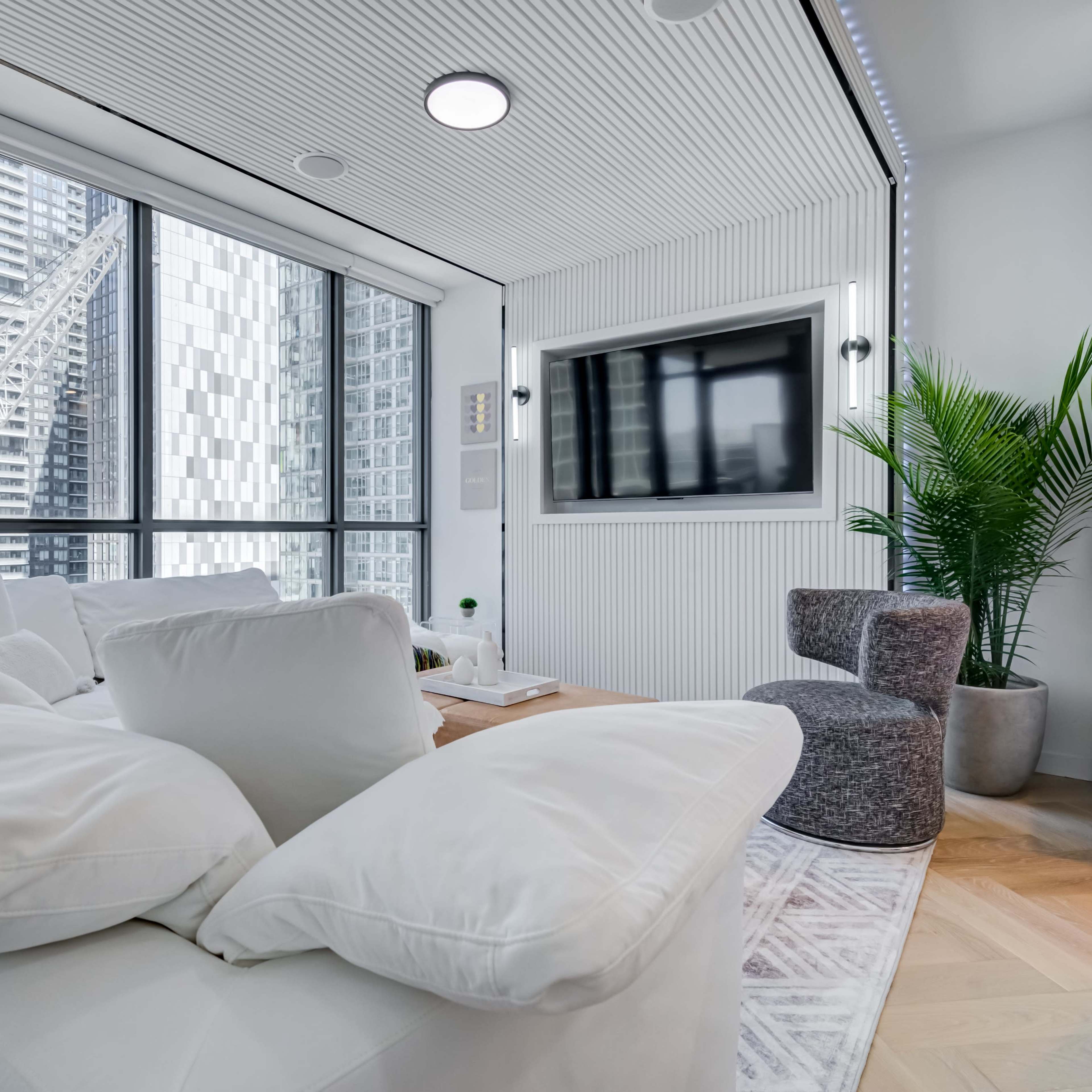 A modern living room with large windows, a sectional sofa, a small armchair, and a television mounted on the wall.