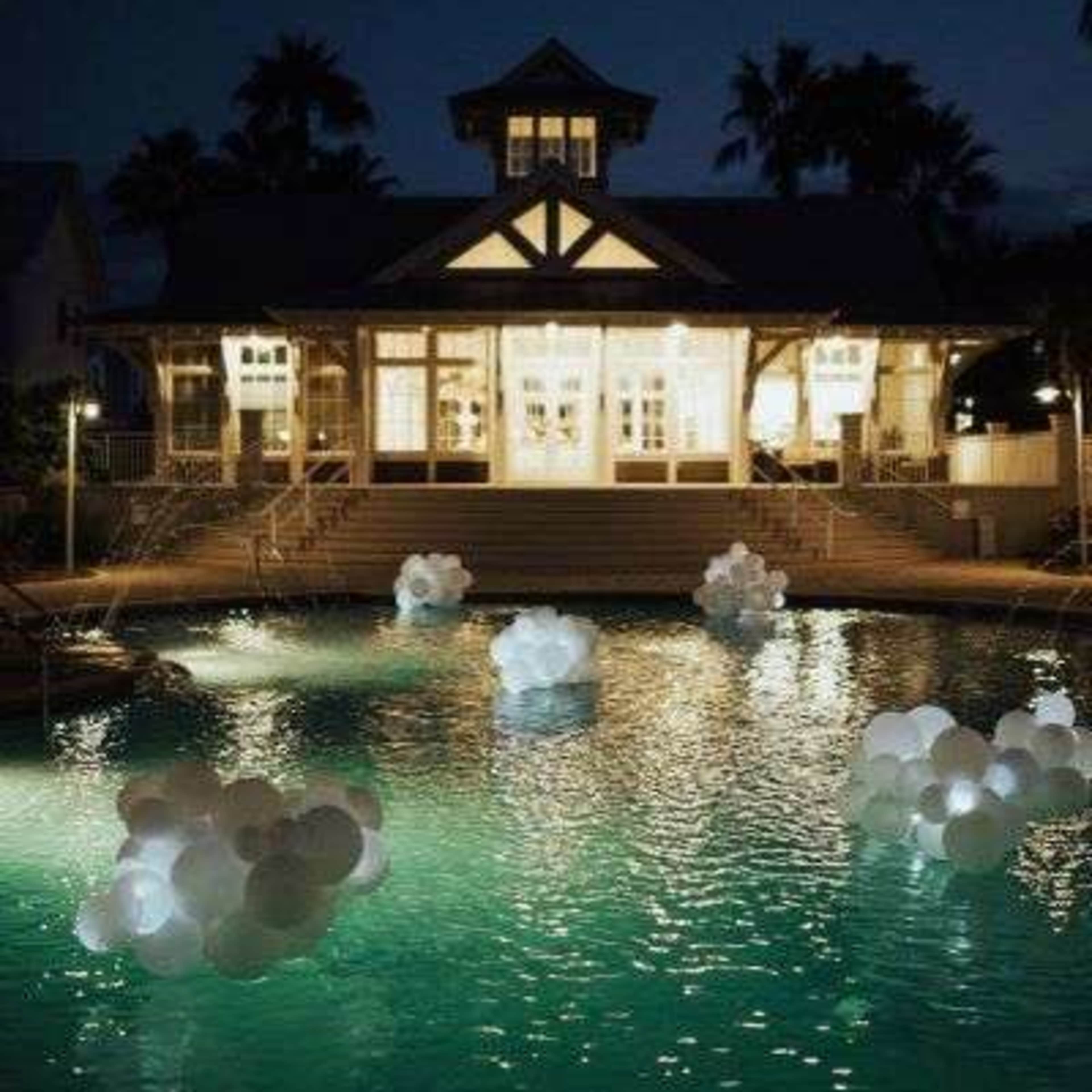 A well-lit house with large windows overlooks a pool featuring illuminated floating decorations at night.