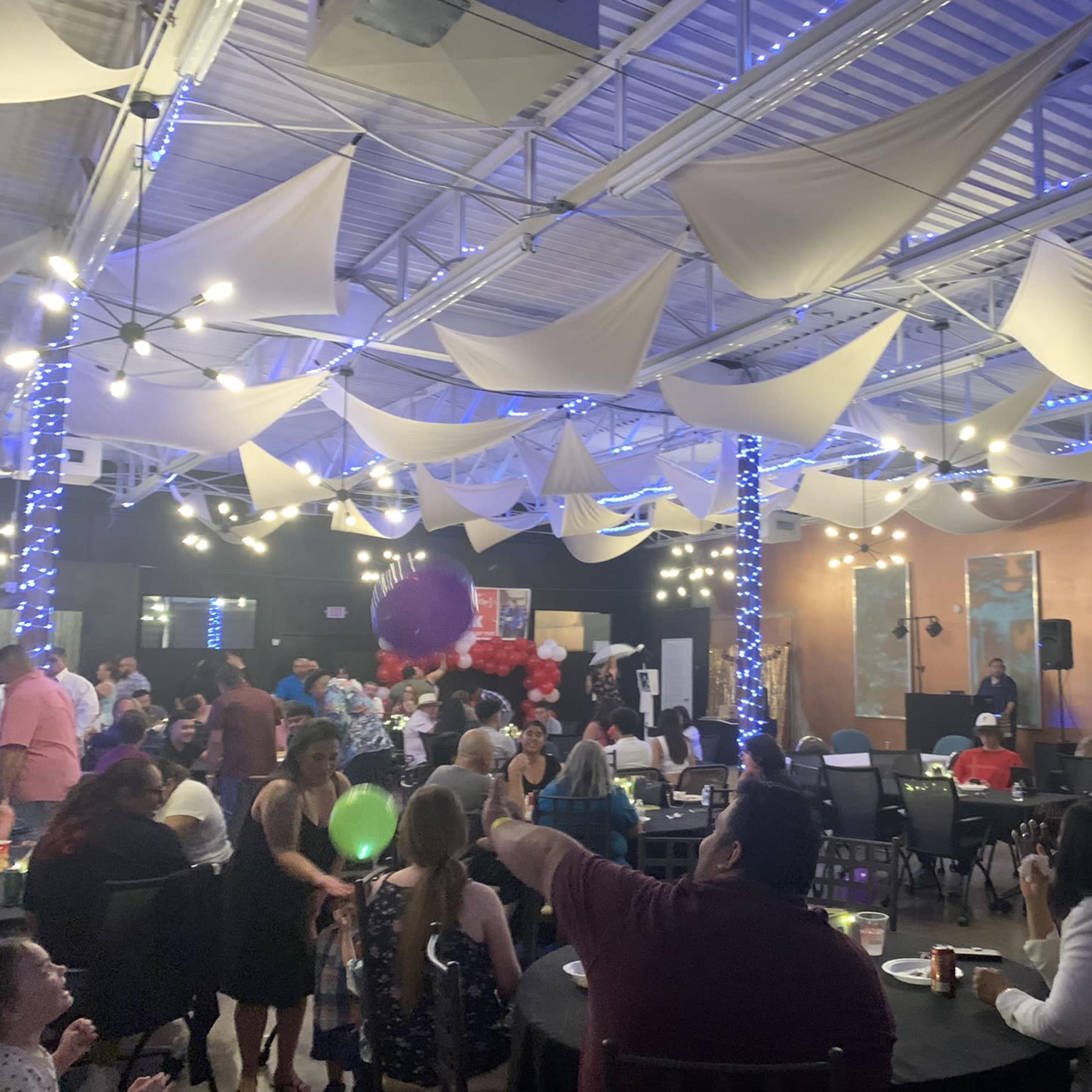 A large indoor space filled with people at tables, decorated with hanging fabric and string lights, hosting an event.