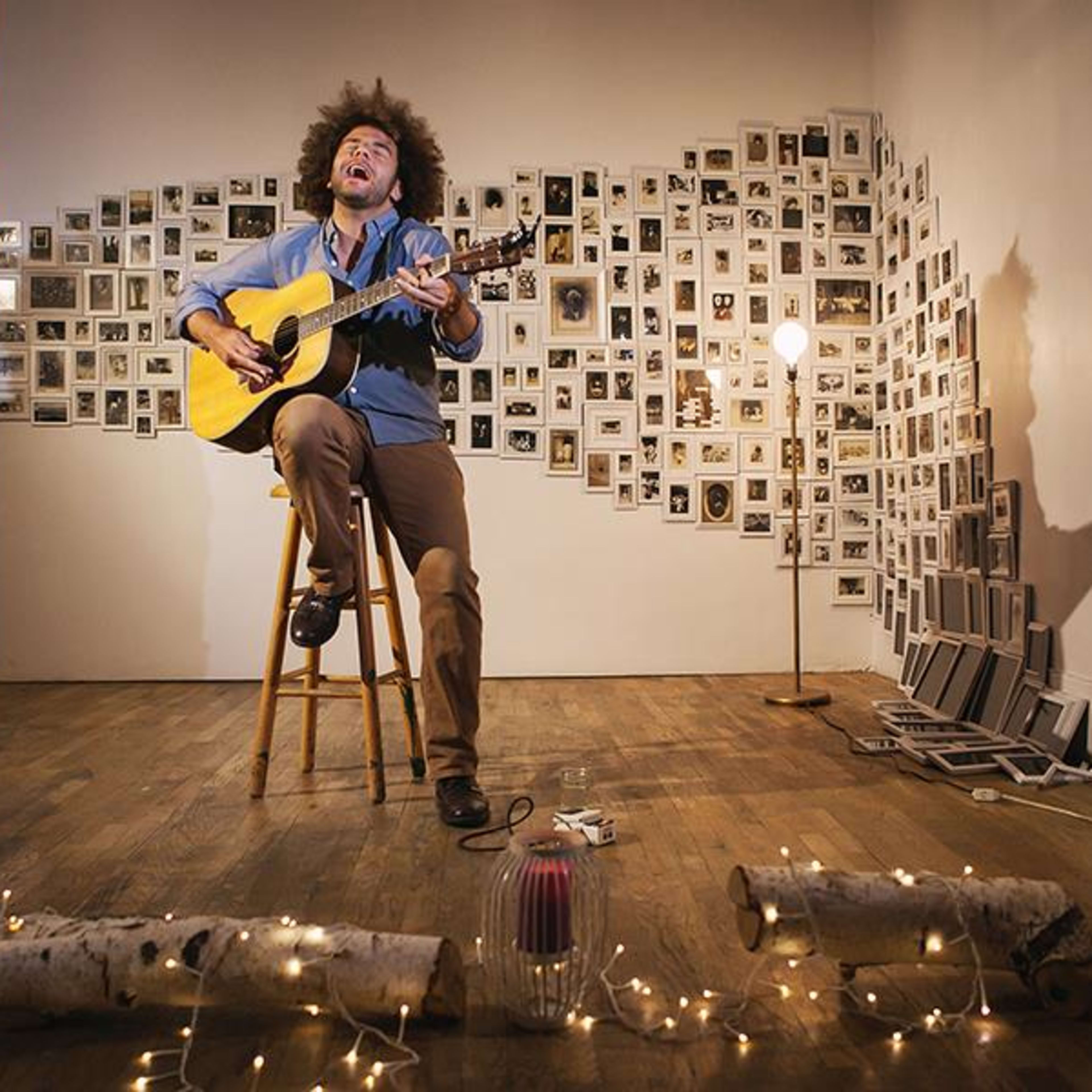 A musician with an afro plays guitar while singing on a stool in a room adorned with numerous framed photographs on the wall.
