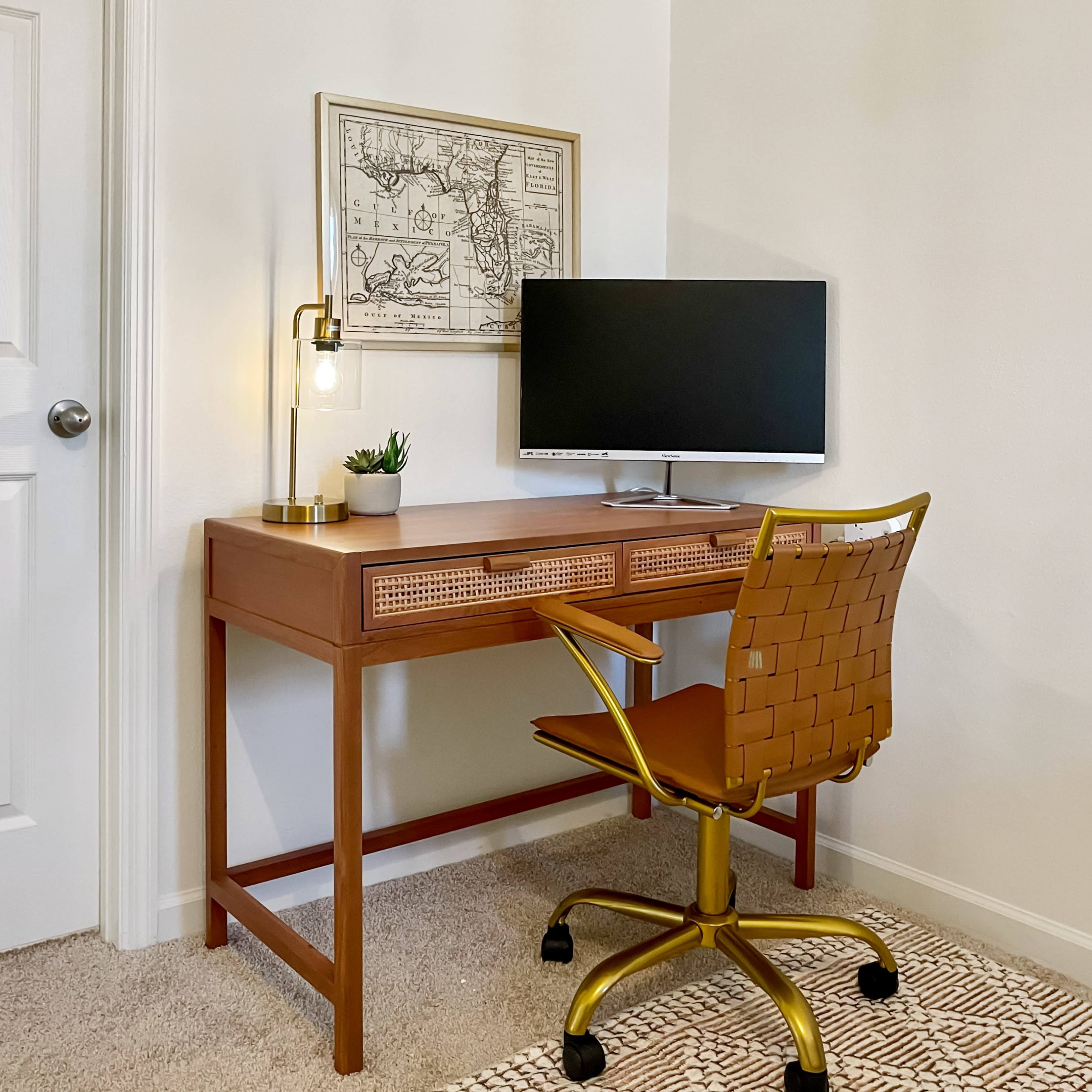 A wooden desk with a computer monitor and a decorative map framed on the wall is situated in a corner of a room, next to a closed door.
