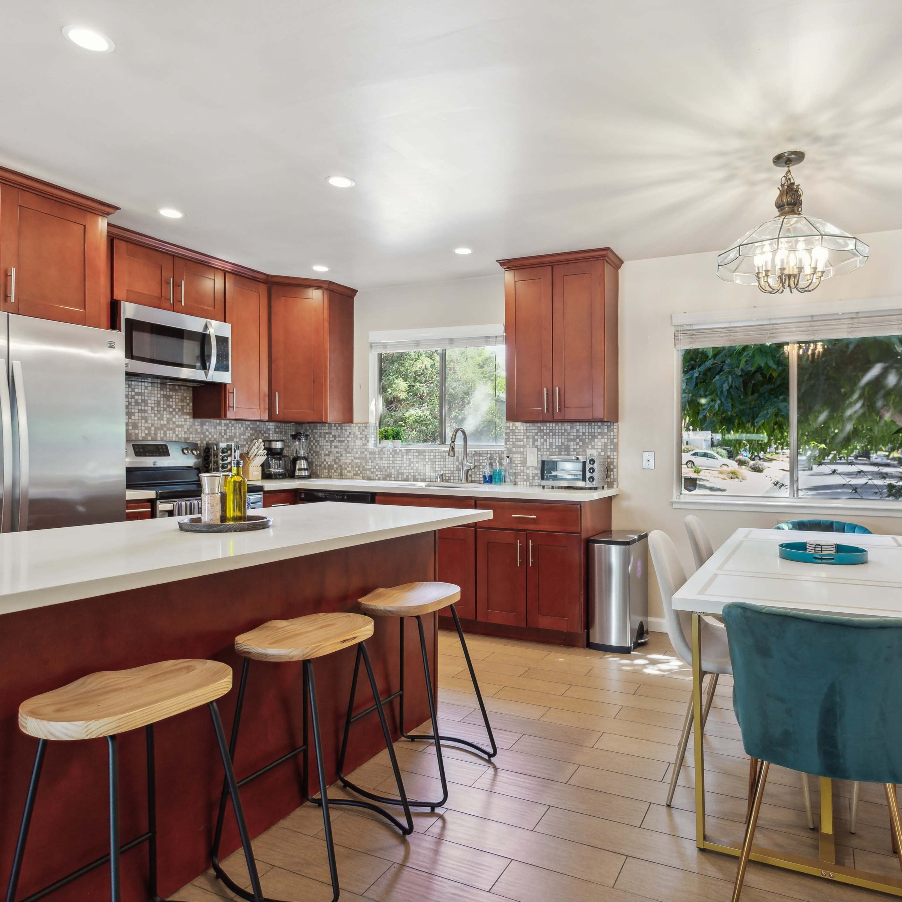 A modern kitchen features wooden cabinets, stainless steel appliances, and a dining area with a table and chairs beside a large window.