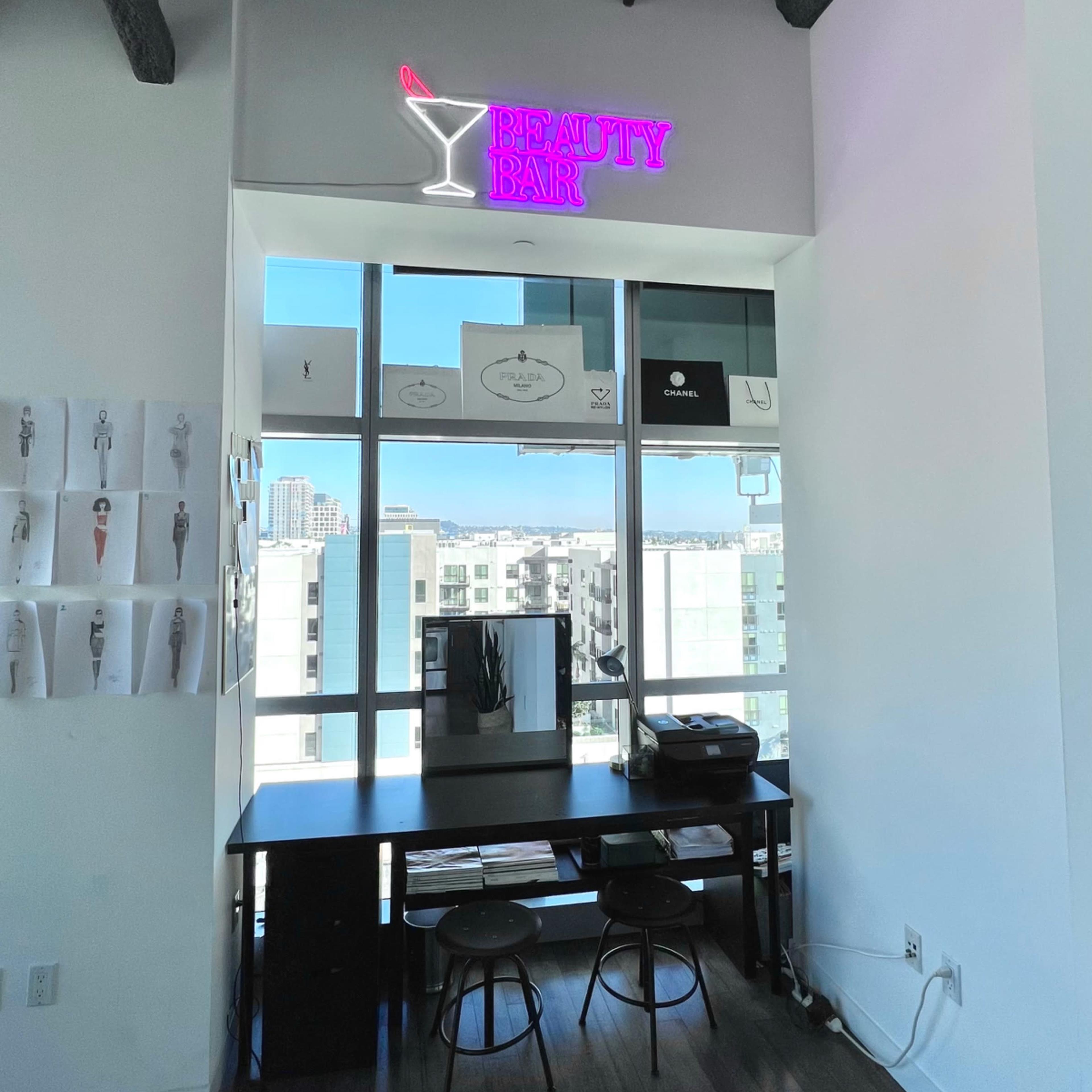 A bright interior with a "BEAUTY BAR" neon sign above a table and two stools near a large window.