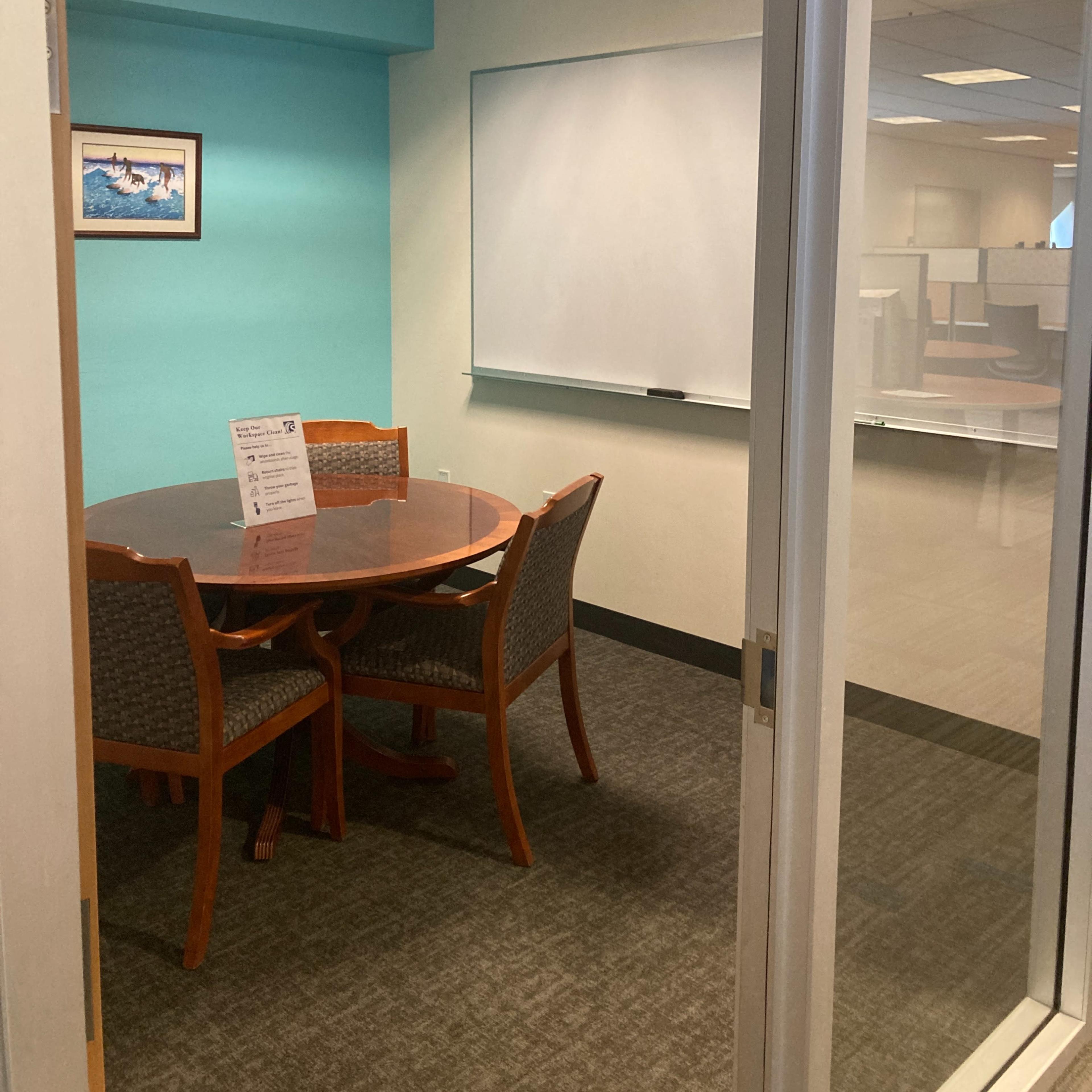 A small conference room featuring a round wooden table surrounded by four chairs, a whiteboard on the wall, and a turquoise-colored wall section.