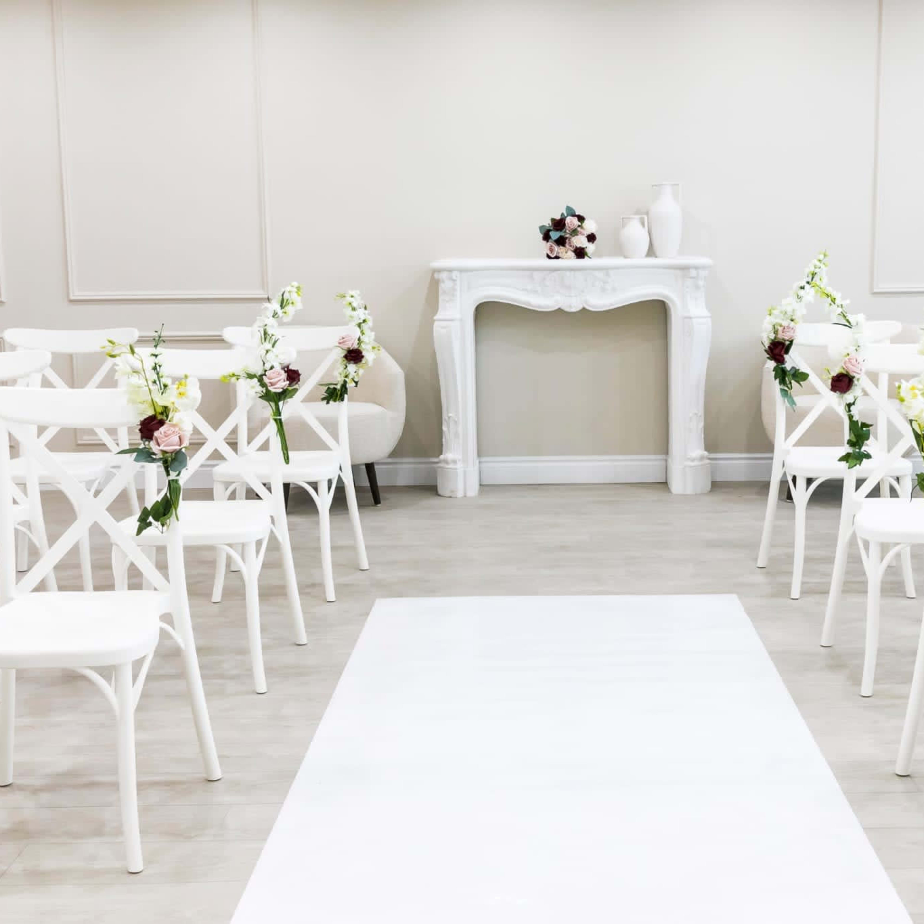 The scene features a minimalist wedding ceremony setup with white chairs arranged in rows facing a central white aisle and a decorative mantelpiece adorned with flowers.