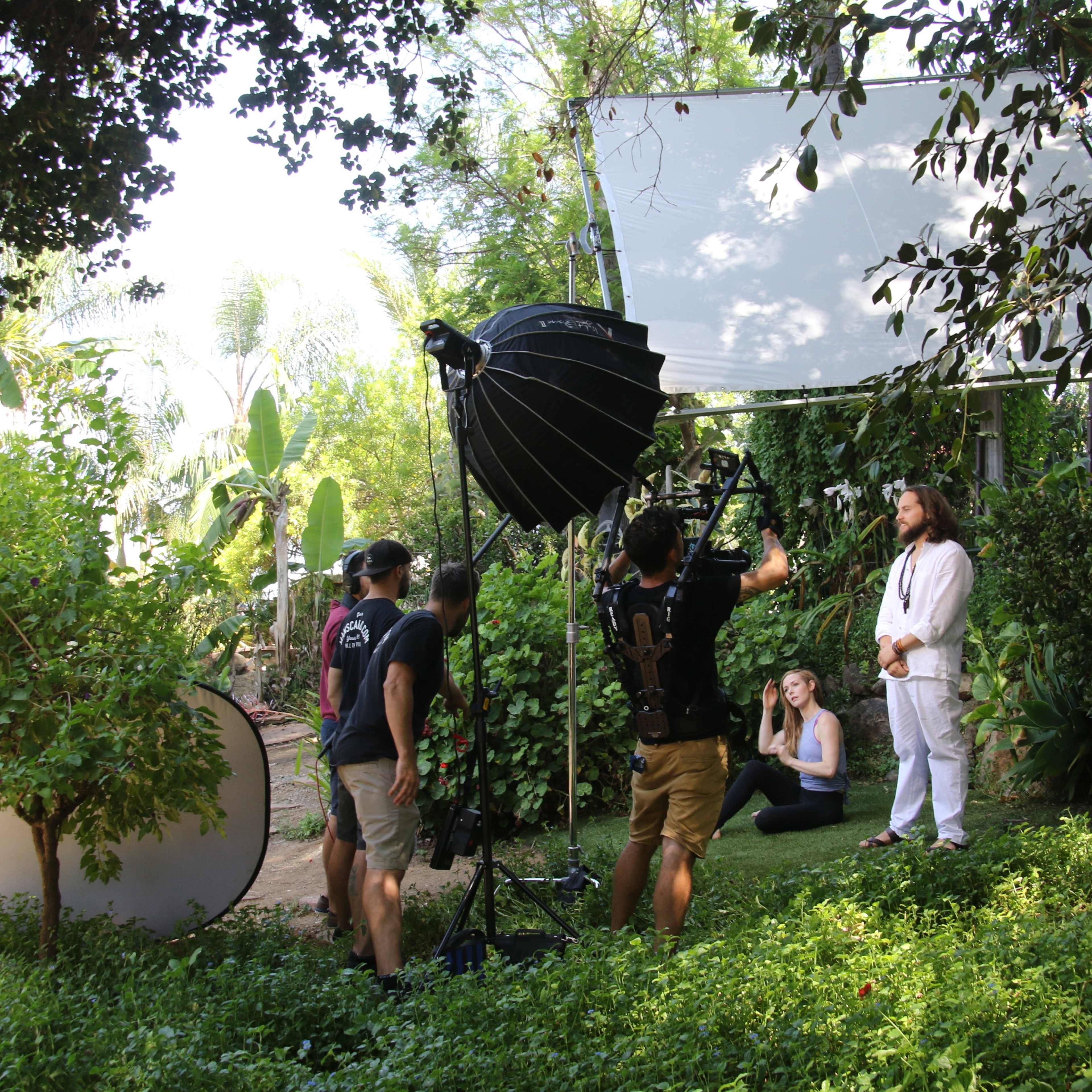 A film crew sets up equipment in a garden to shoot a scene featuring a man in white clothing standing among greenery while others prepare and observe.