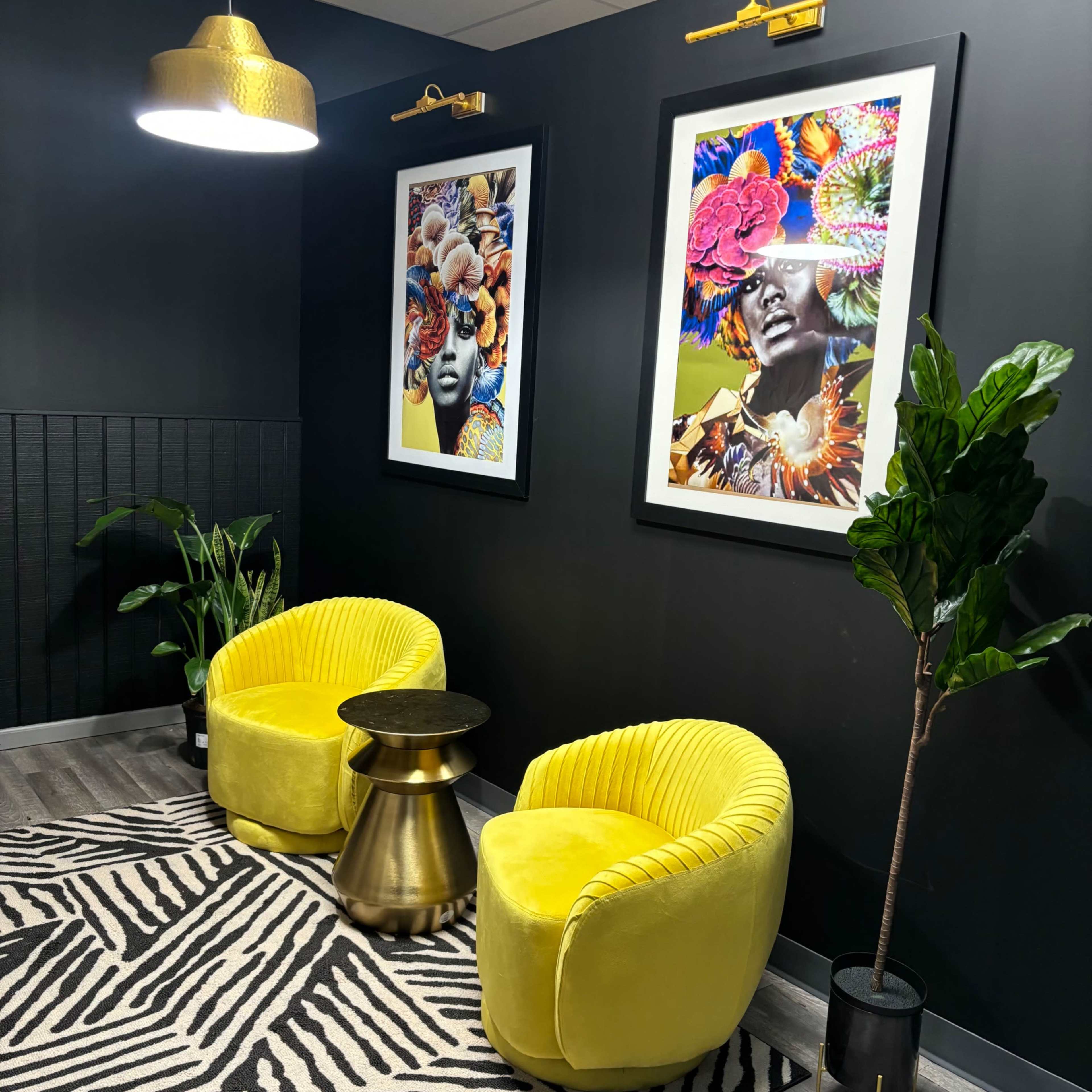 The scene features two yellow armchairs flanking a round gold table, set against a dark wall adorned with vibrant framed artwork and complemented by a small potted plant.