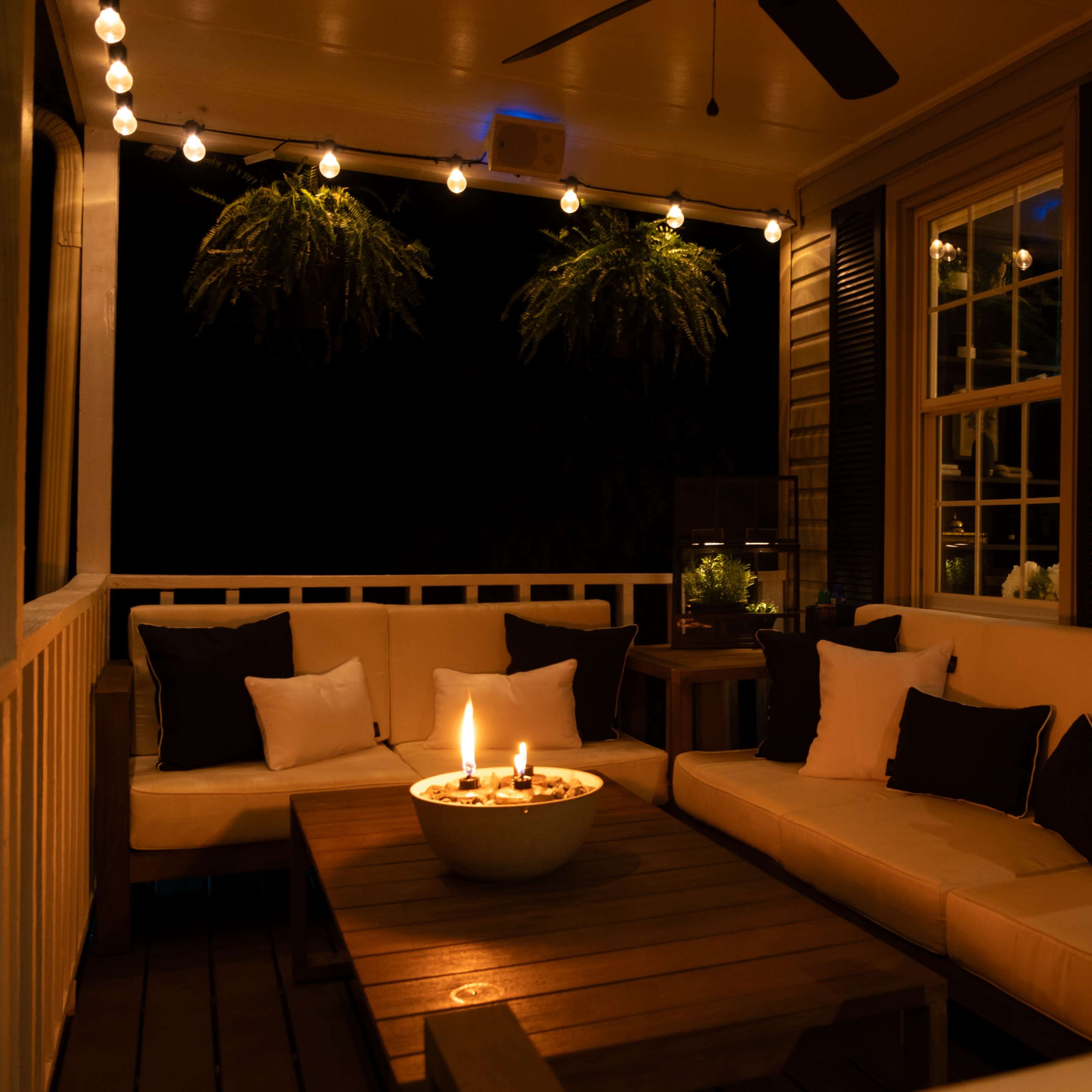 A cozy porch area features a seating arrangement with a fire pit at the center, surrounded by string lights and lush greenery.