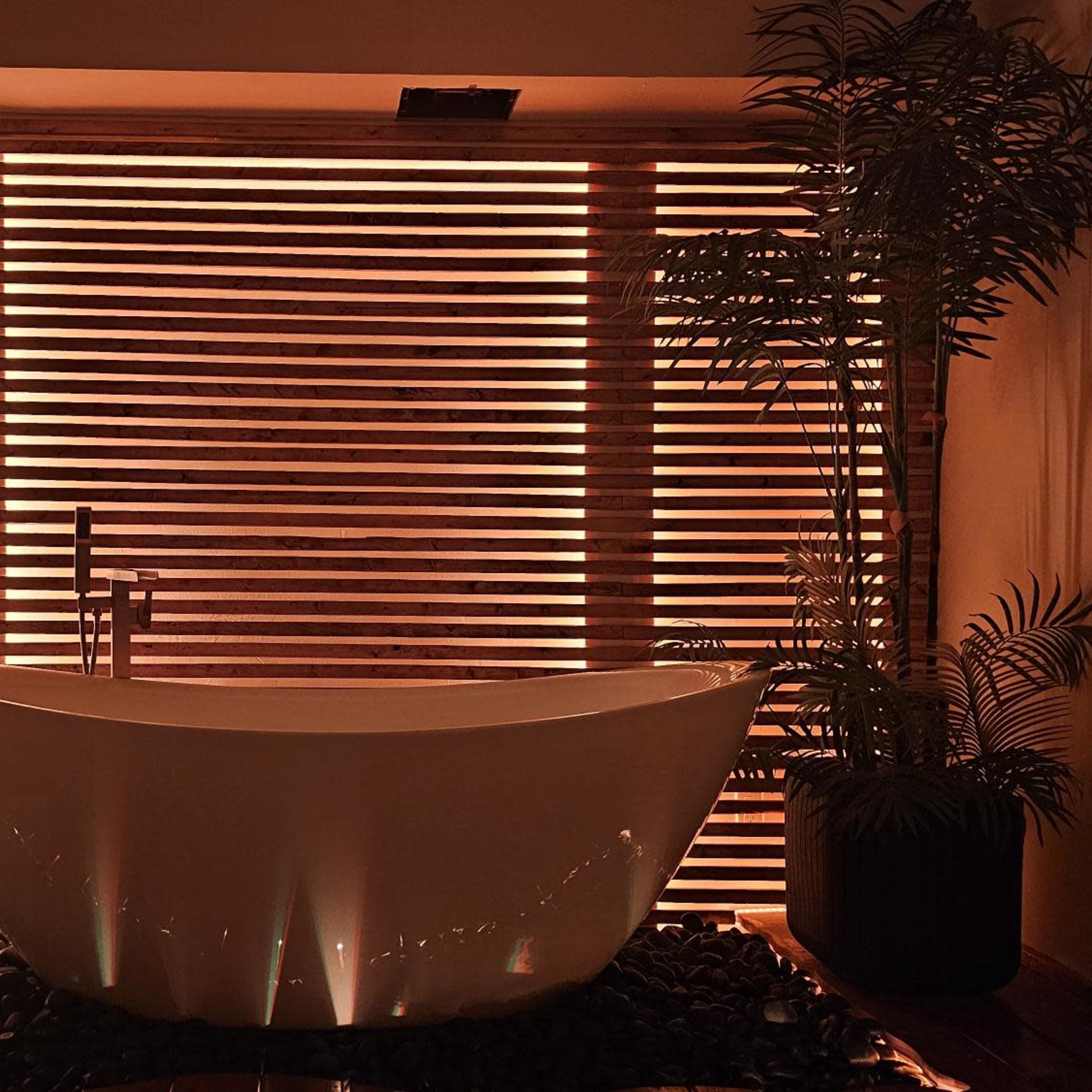A white freestanding bathtub is positioned in front of a slatted wooden screen illuminated by warm light, surrounded by potted plants and dark stones on the floor.