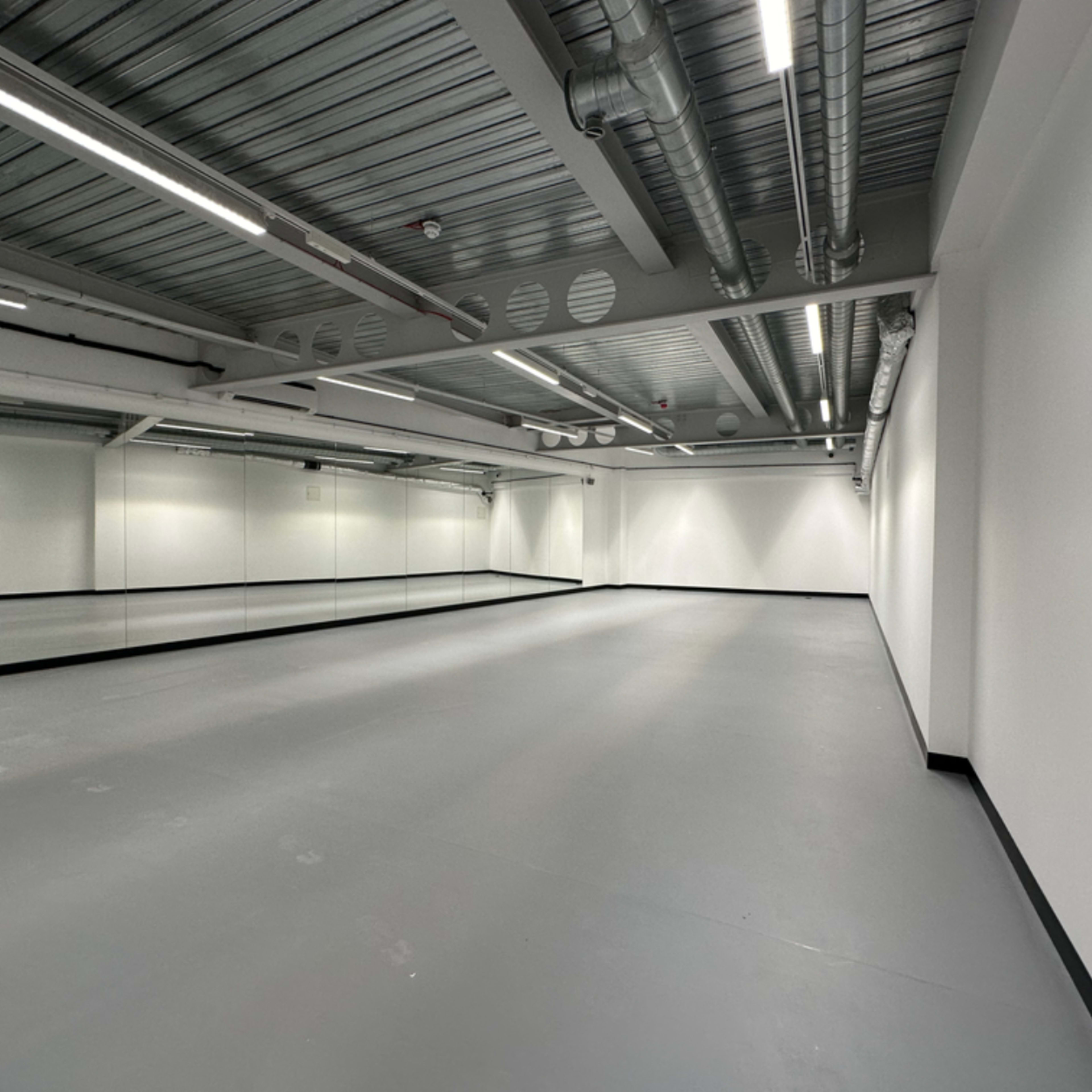 A spacious, empty room with gray flooring and a wall of mirrors under exposed ceiling infrastructure.