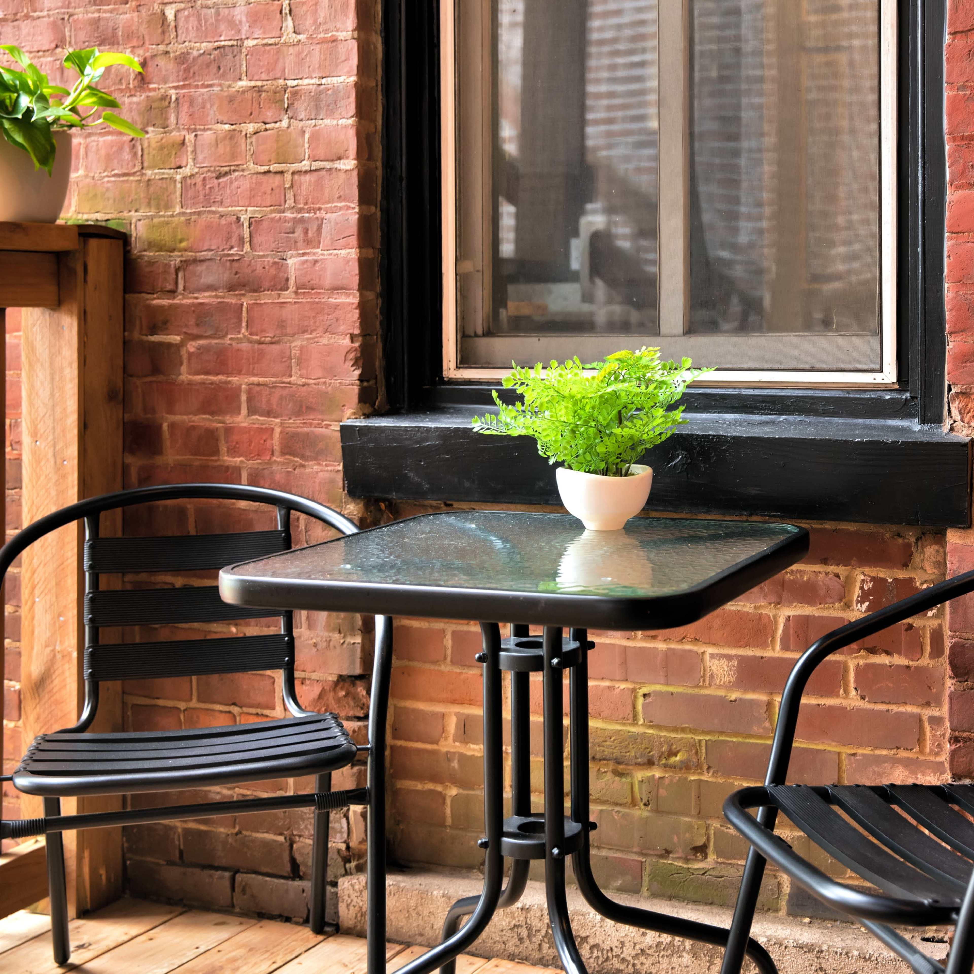 A small balcony features a glass table with two black chairs and a potted plant on the table, situated against a brick wall.