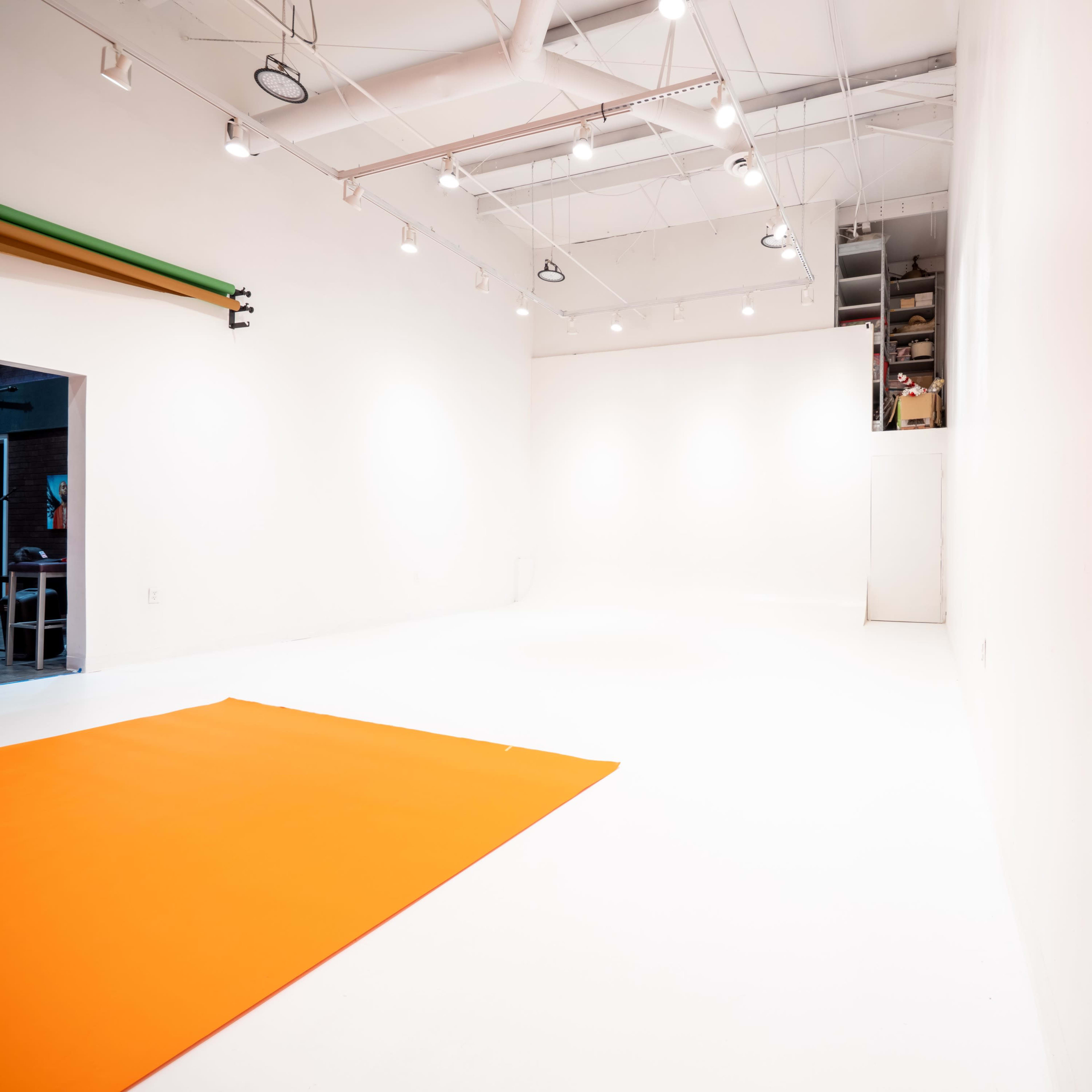 A large, empty studio space features white walls, a white floor, and an orange mat in one corner.