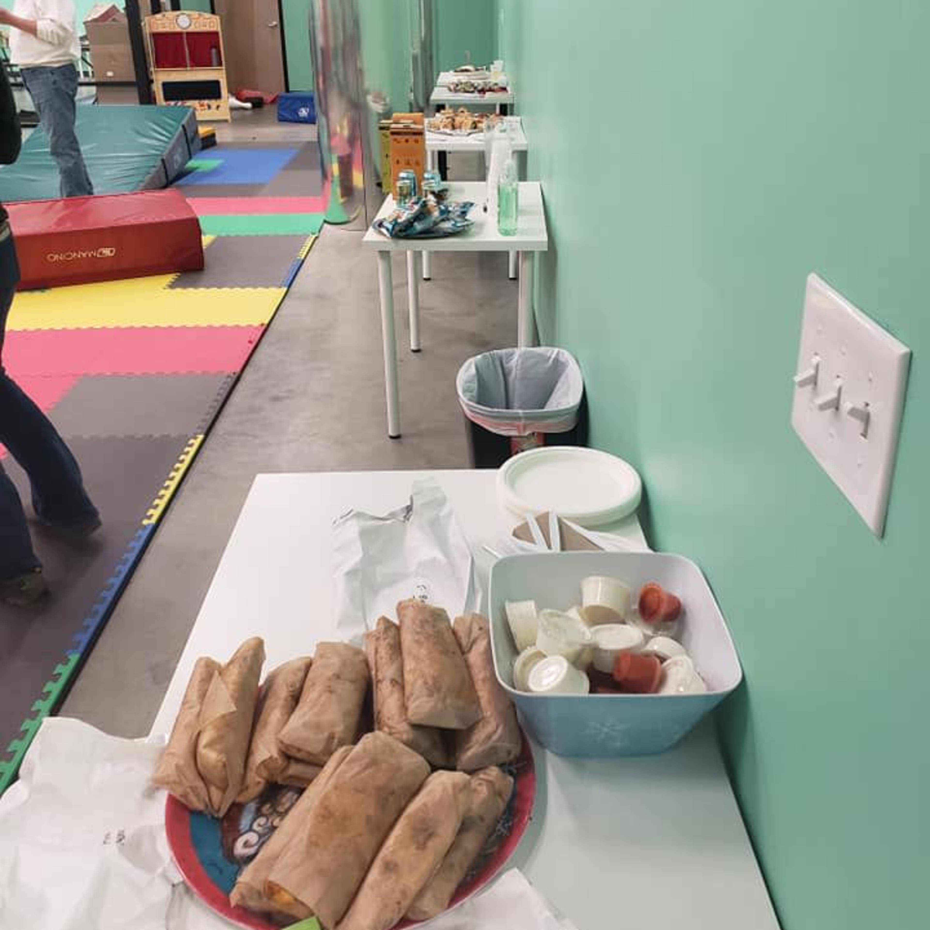 A green-walled room with a table displaying food items, including wrapped burritos and condiments, alongside a play area visible in the background.