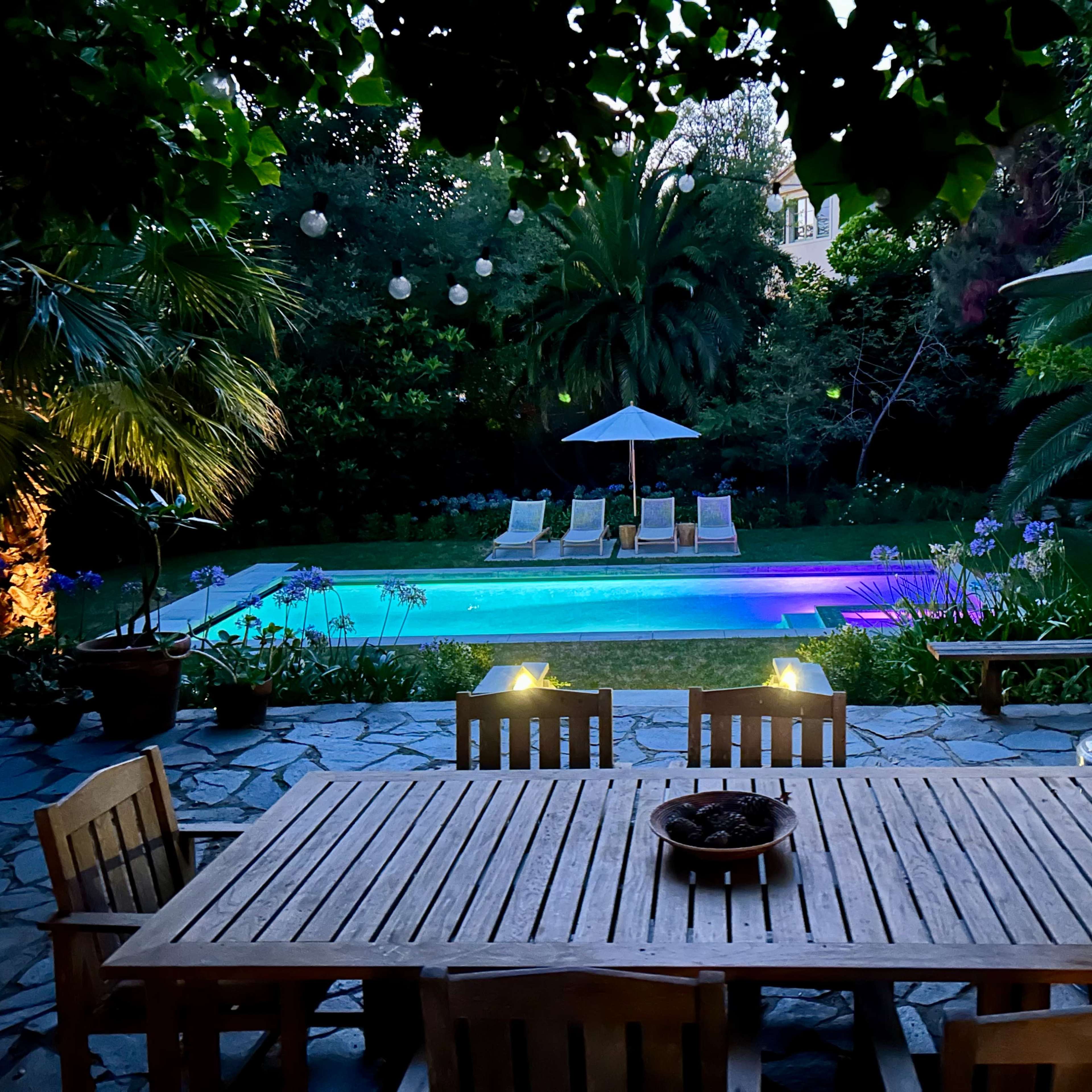 A wooden dining table is set in the foreground, overlooking a lit pool area surrounded by palm trees and lounge chairs.