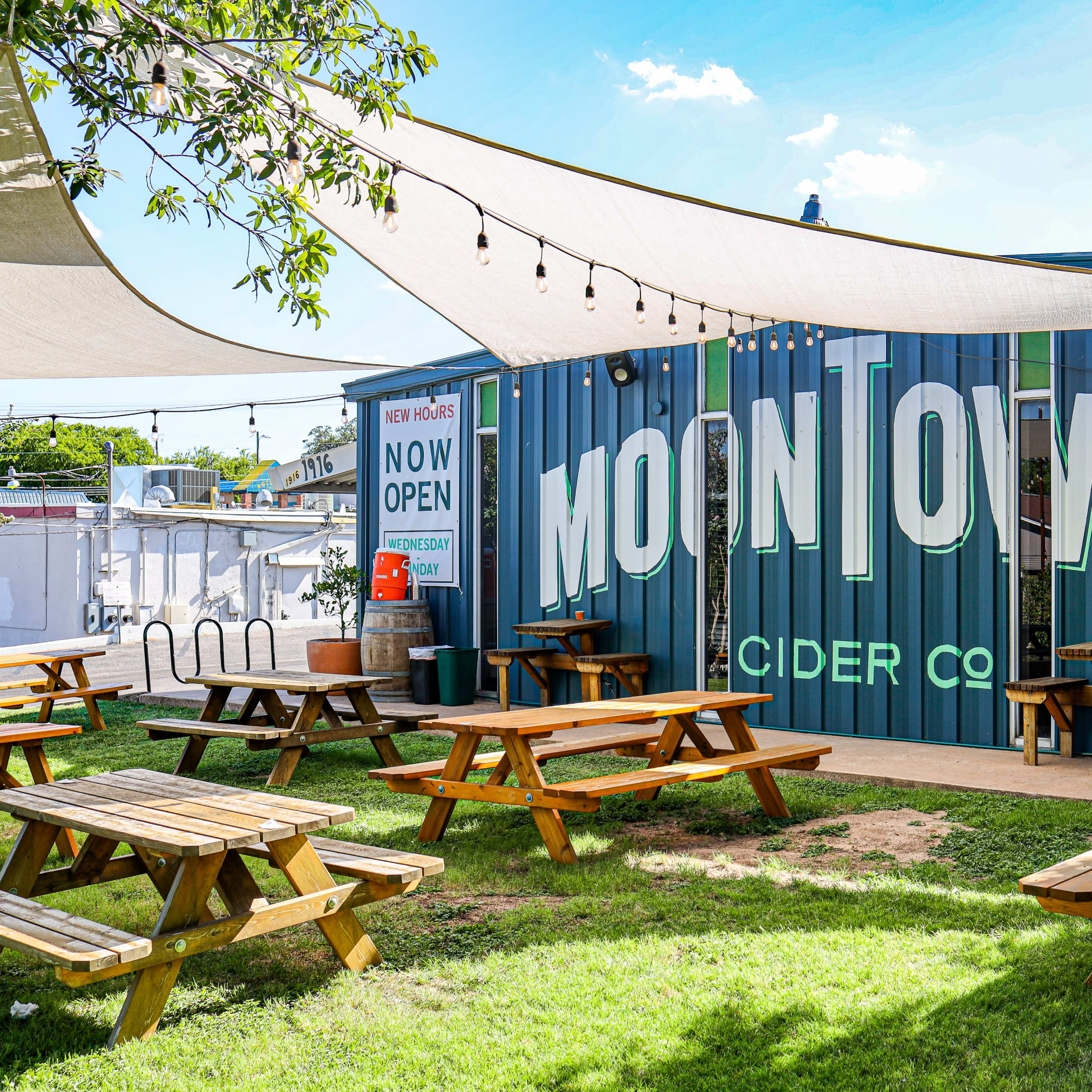 A cider company with a large mural on its side and outdoor seating made up of wooden picnic tables under shaded canopies.