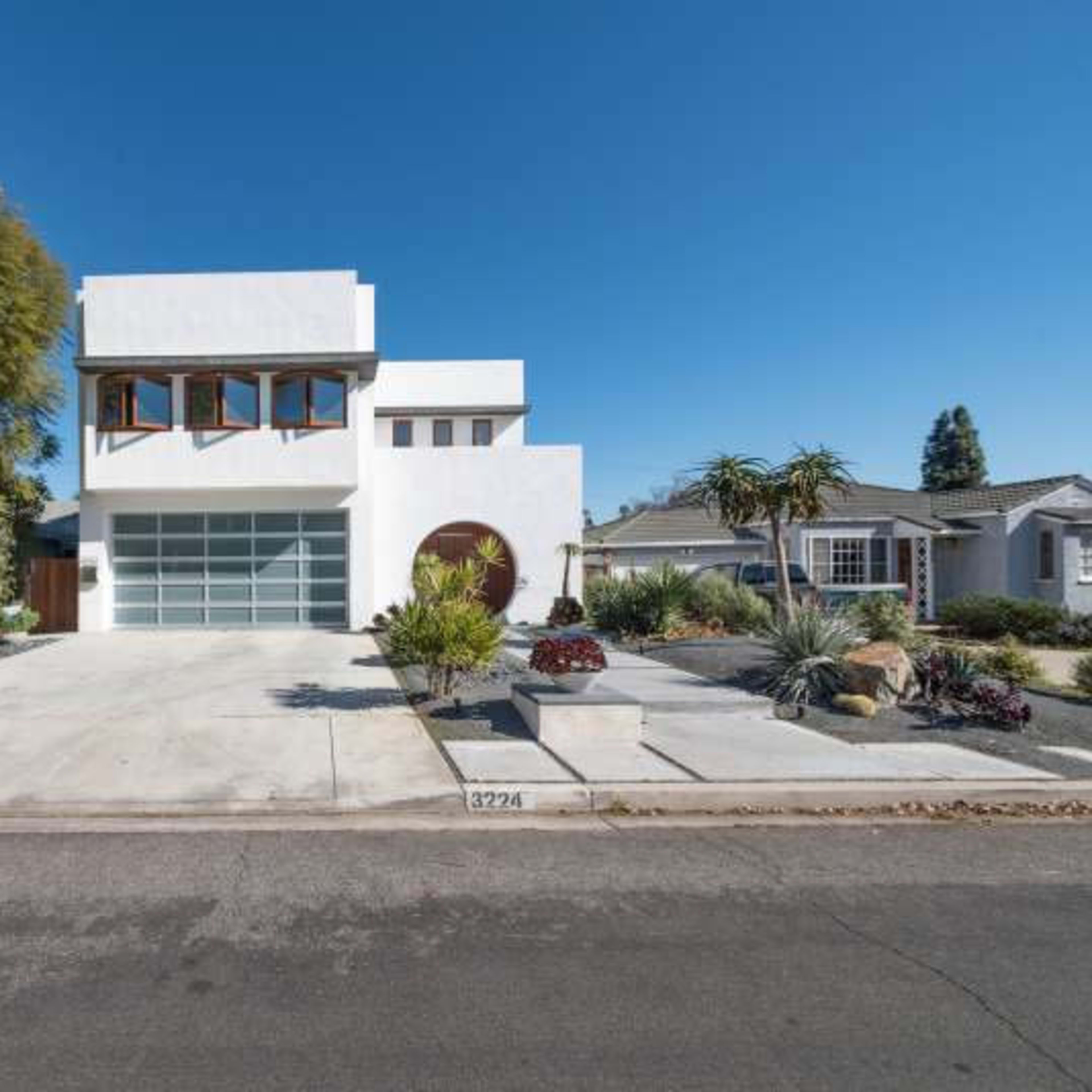 A modern two-story white house with large windows and a spacious front driveway, surrounded by landscaped greenery.