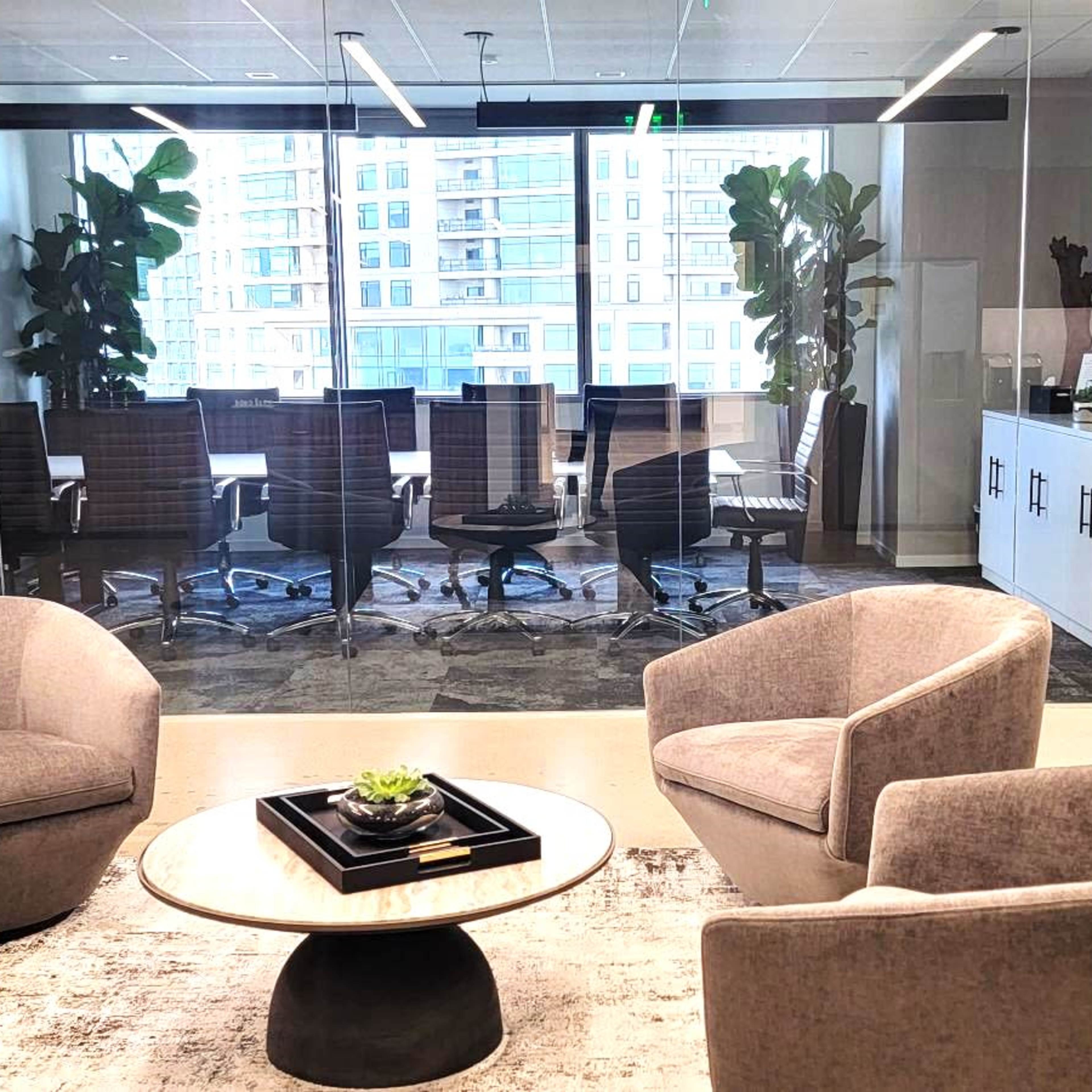 A modern office space features a seating area with three upholstered chairs and a round coffee table, overlooking a conference room with a large window and several office chairs.