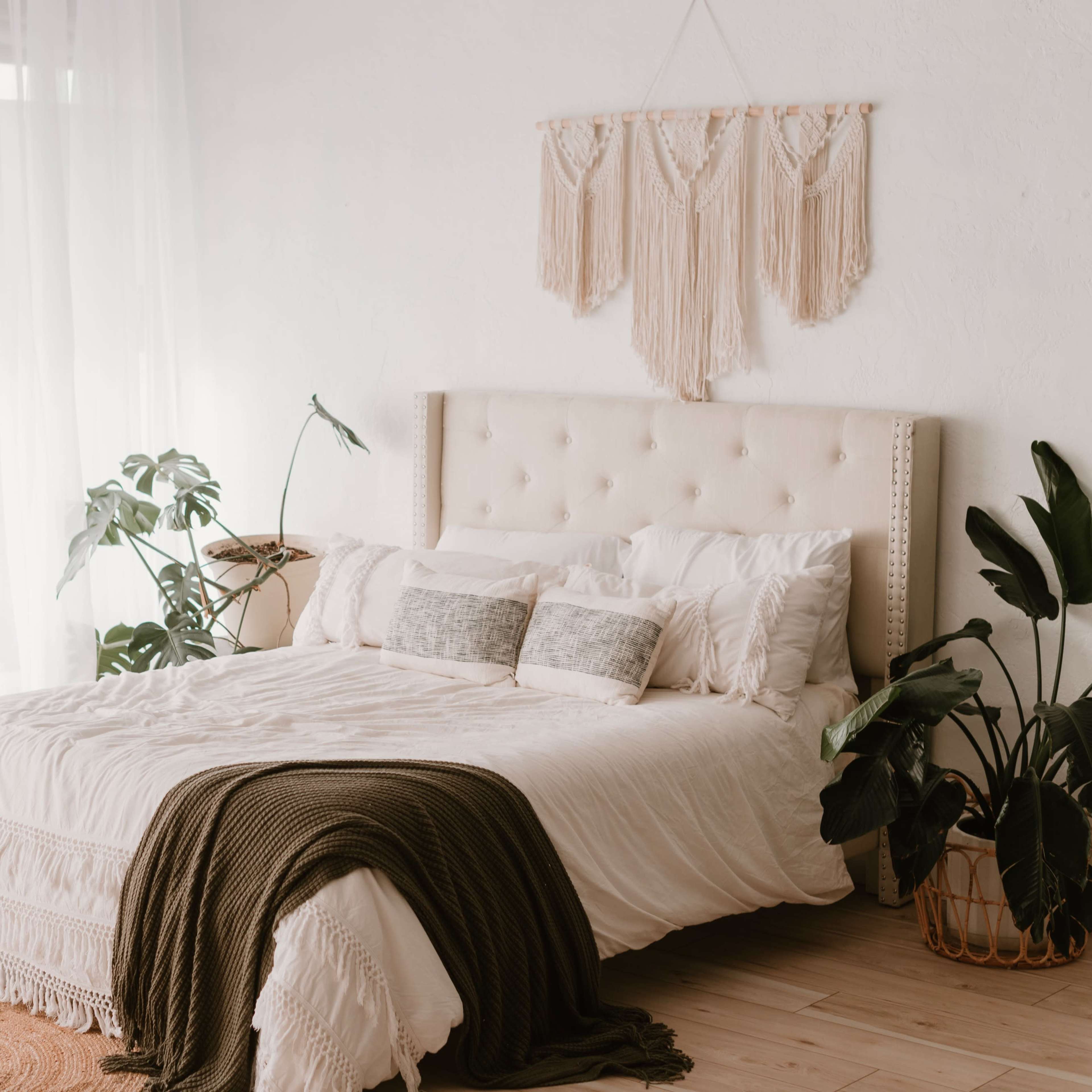 A neatly made bed with a decorative headboard, surrounded by indoor plants and bathed in natural light from large sheer curtains.