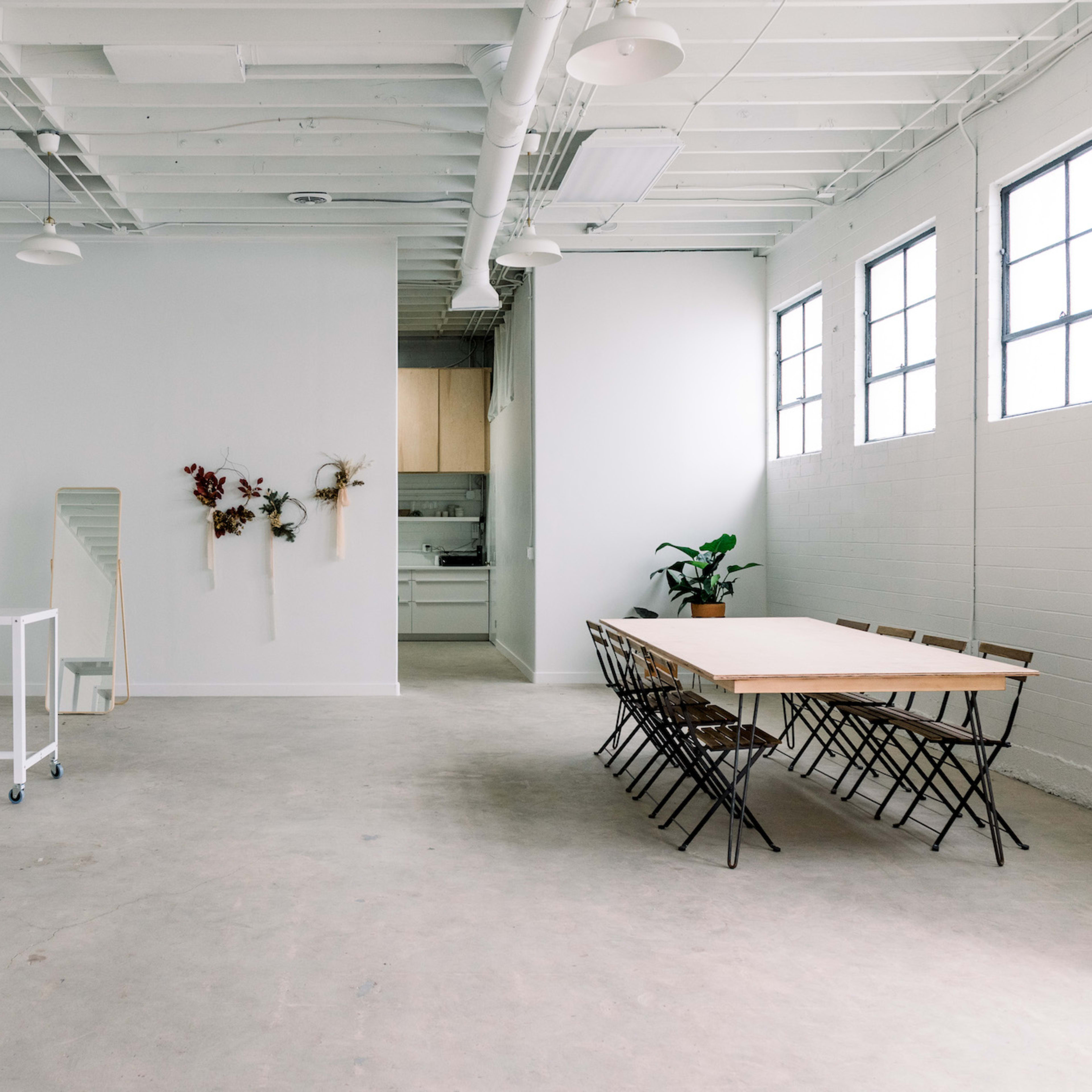A spacious, minimalistic room with concrete flooring, large windows, a long wooden table with chairs, and a kitchenette in the background.
