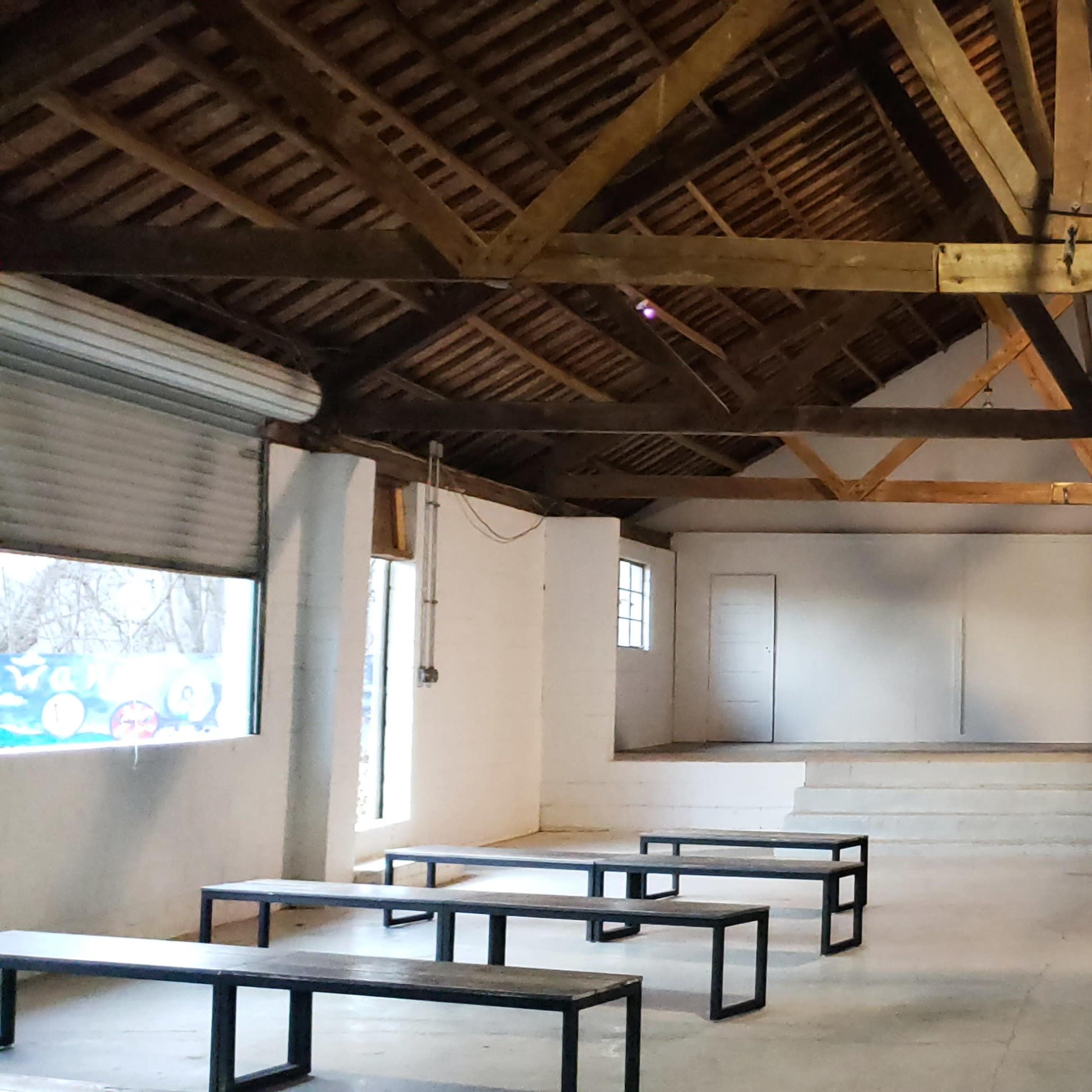 A spacious indoor venue with a high wooden ceiling, featuring benches arranged in front of a raised stage and two doors on the side walls.