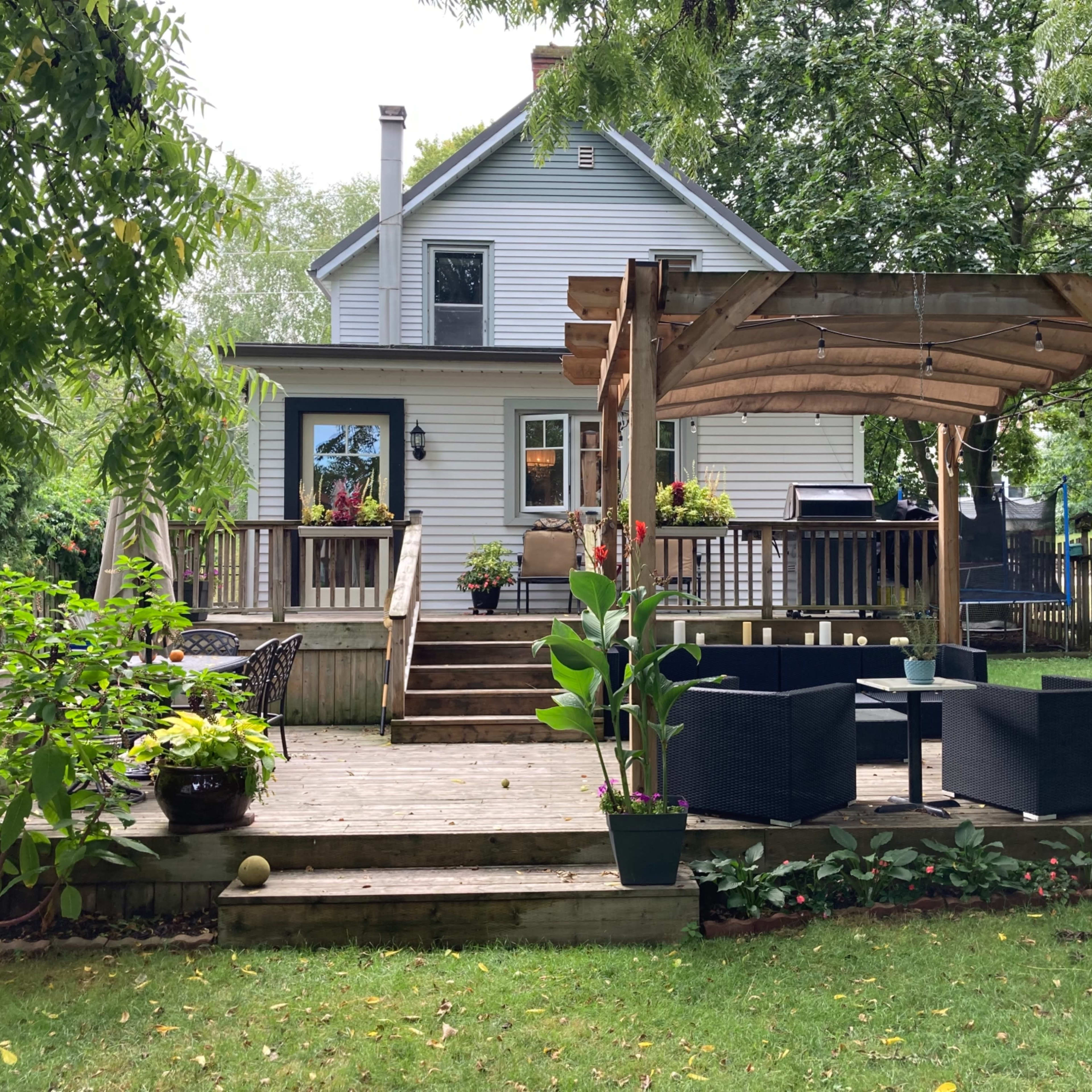 A backyard with a wooden deck, a pergola, and seating area, adjacent to a two-story white house surrounded by trees and plants.