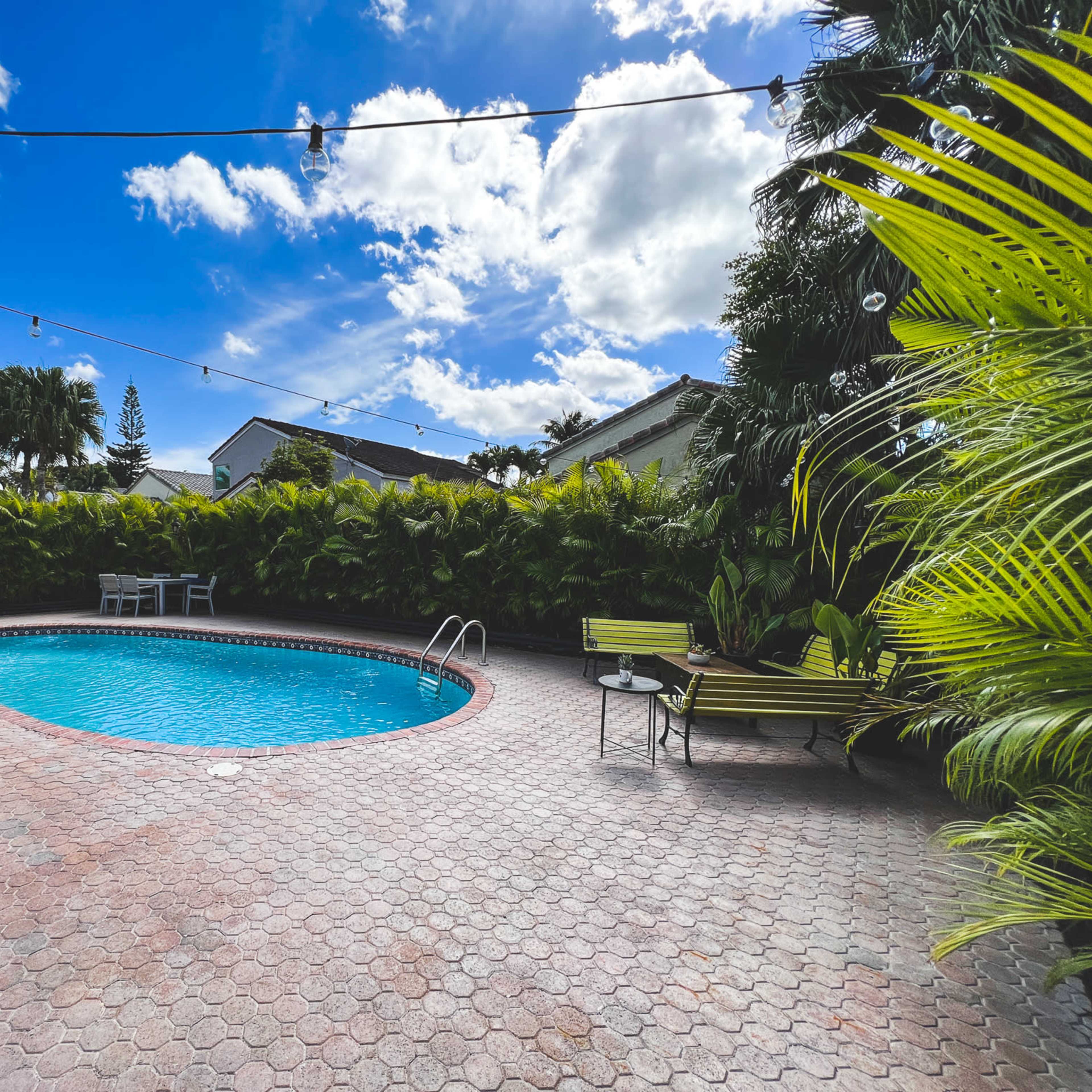 A swimming pool surrounded by a paved deck, with lounge chairs and lush greenery in the background under a partly cloudy sky.