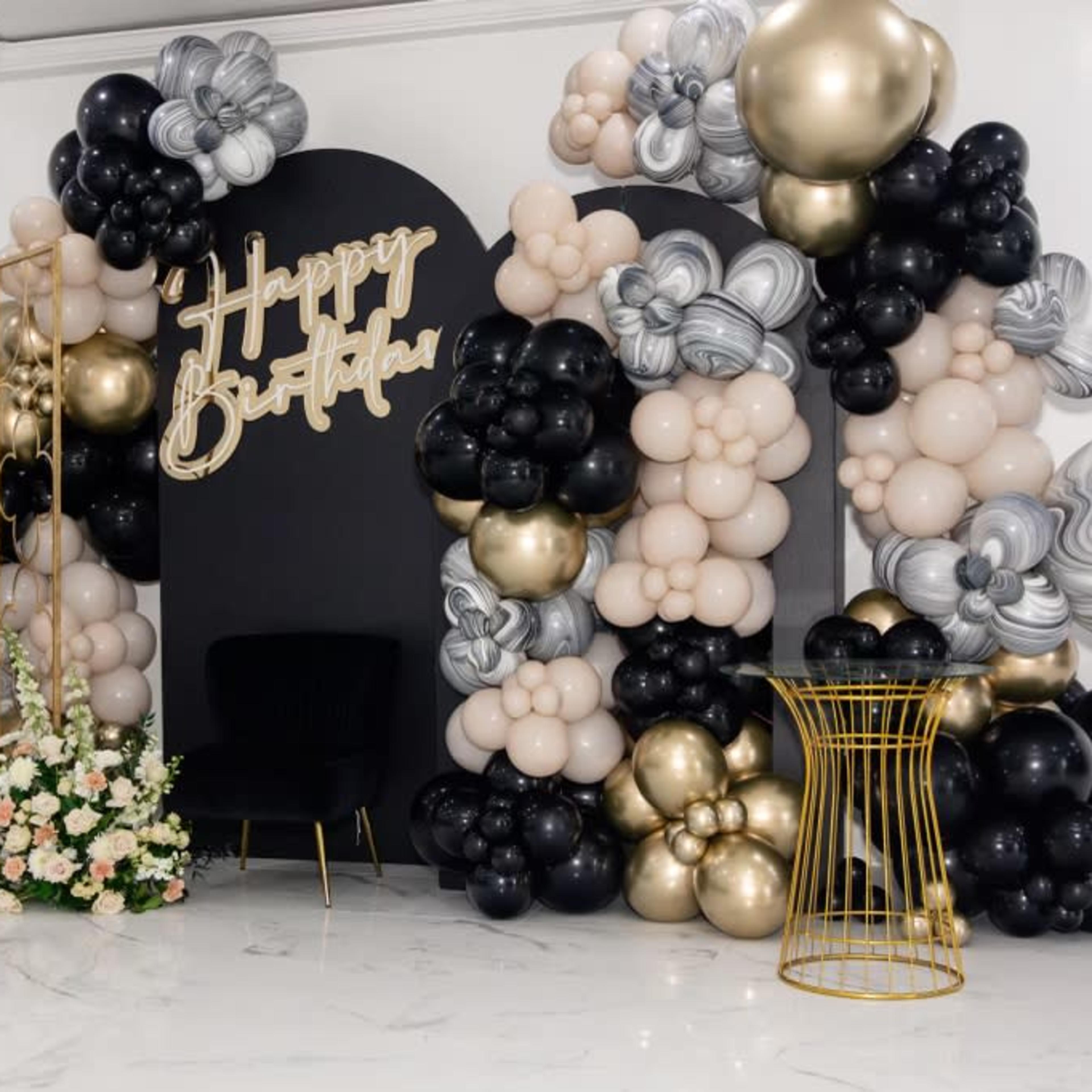 The scene features a decorated birthday backdrop with black, gold, and marble-patterned balloons, along with floral arrangements and a black seating area.