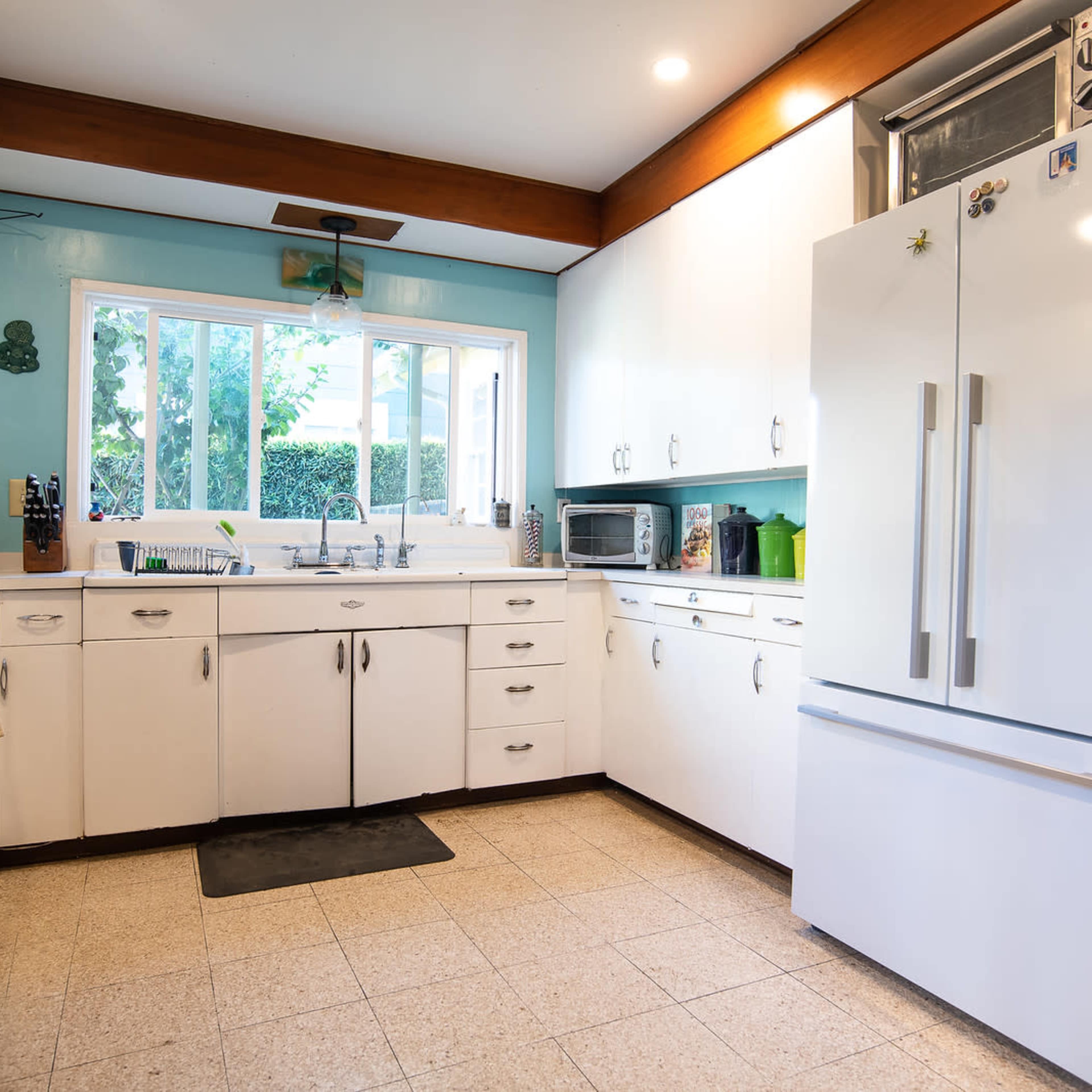 A bright kitchen with white cabinets, a large refrigerator, and a window overlooking an outdoor area.