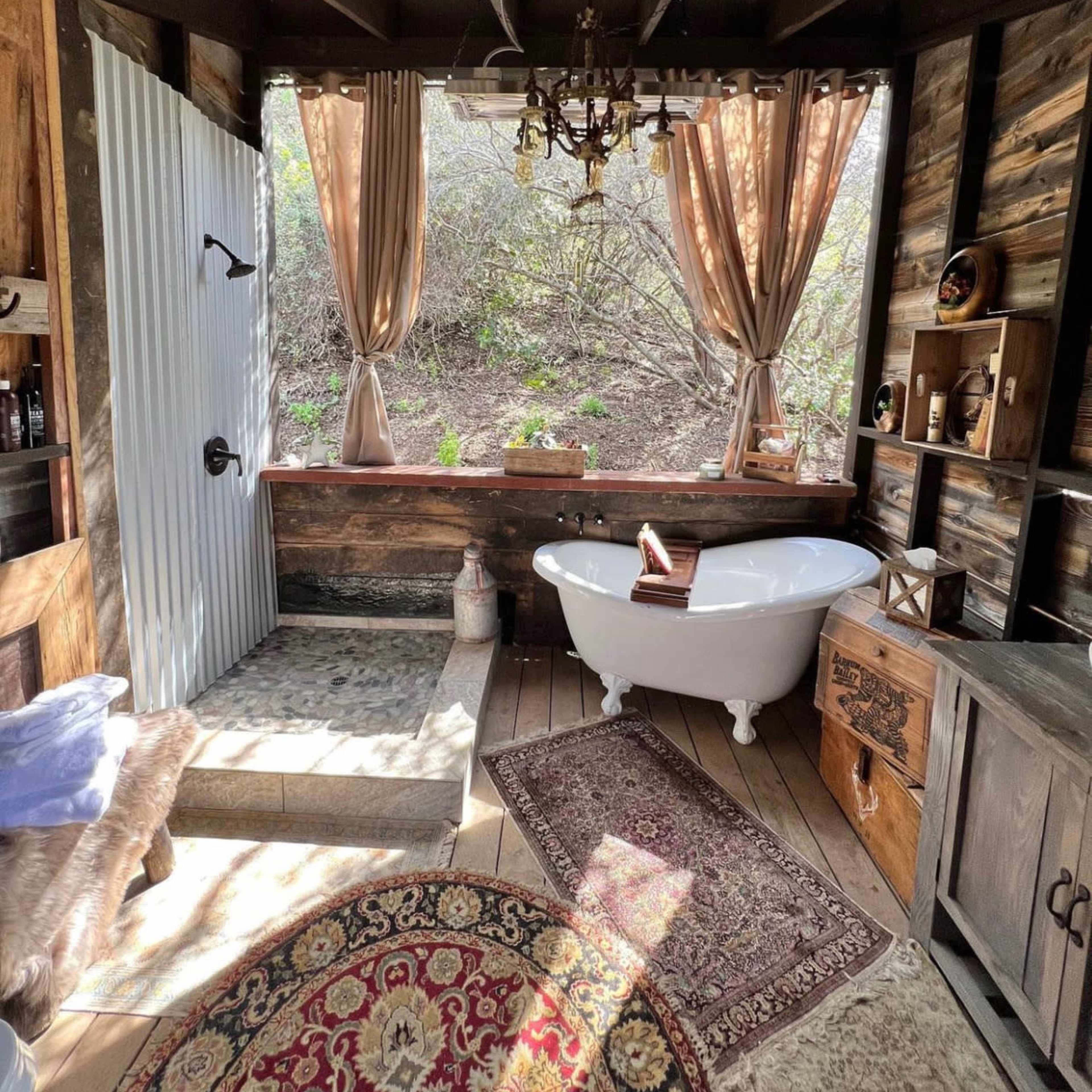 A rustic bathroom featuring a freestanding bathtub, a vintage chandelier, wooden walls, and large windows framed by curtains.