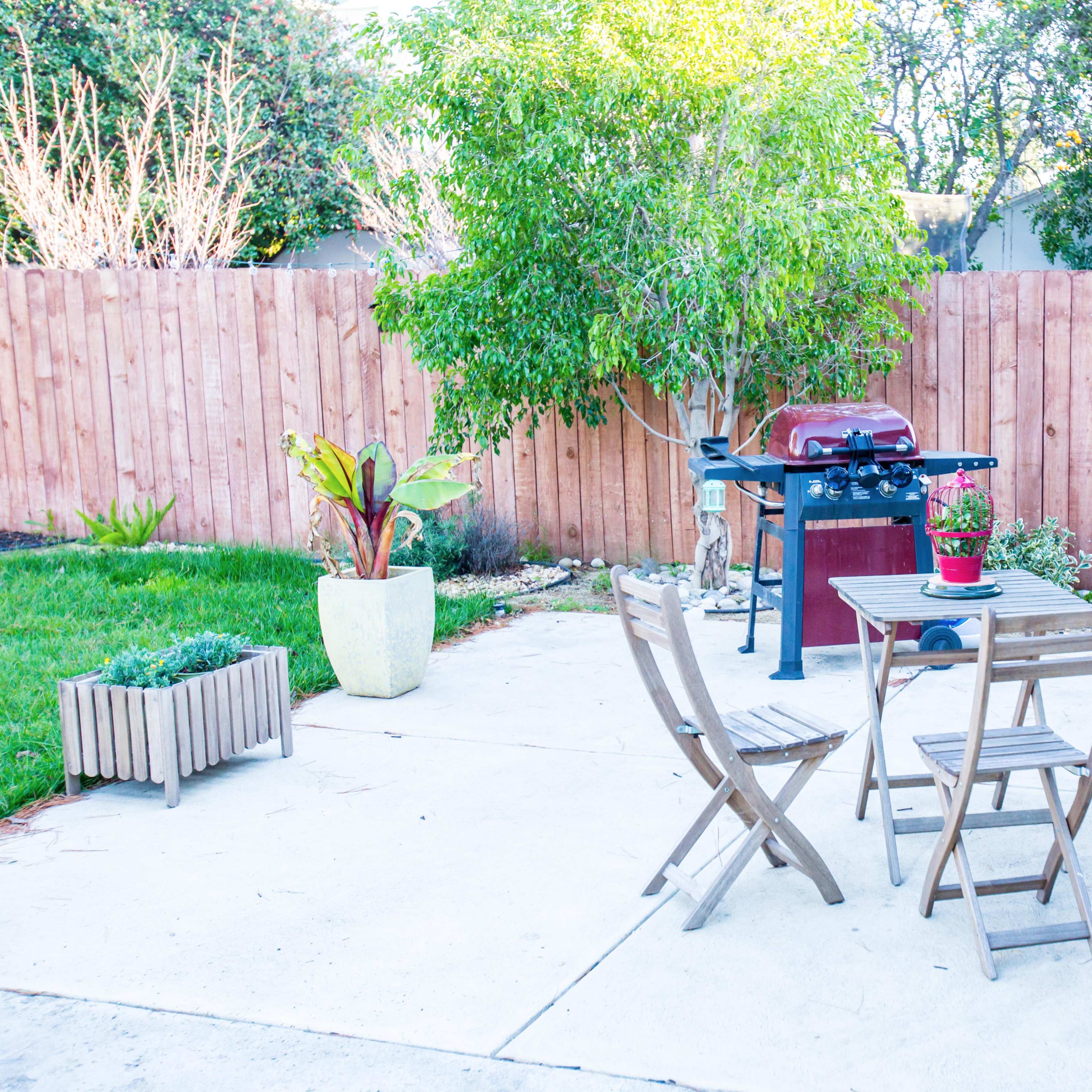 A small patio features a table and two chairs, a barbecue grill, a potted plant, and an air conditioning unit surrounded by a wooden fence.