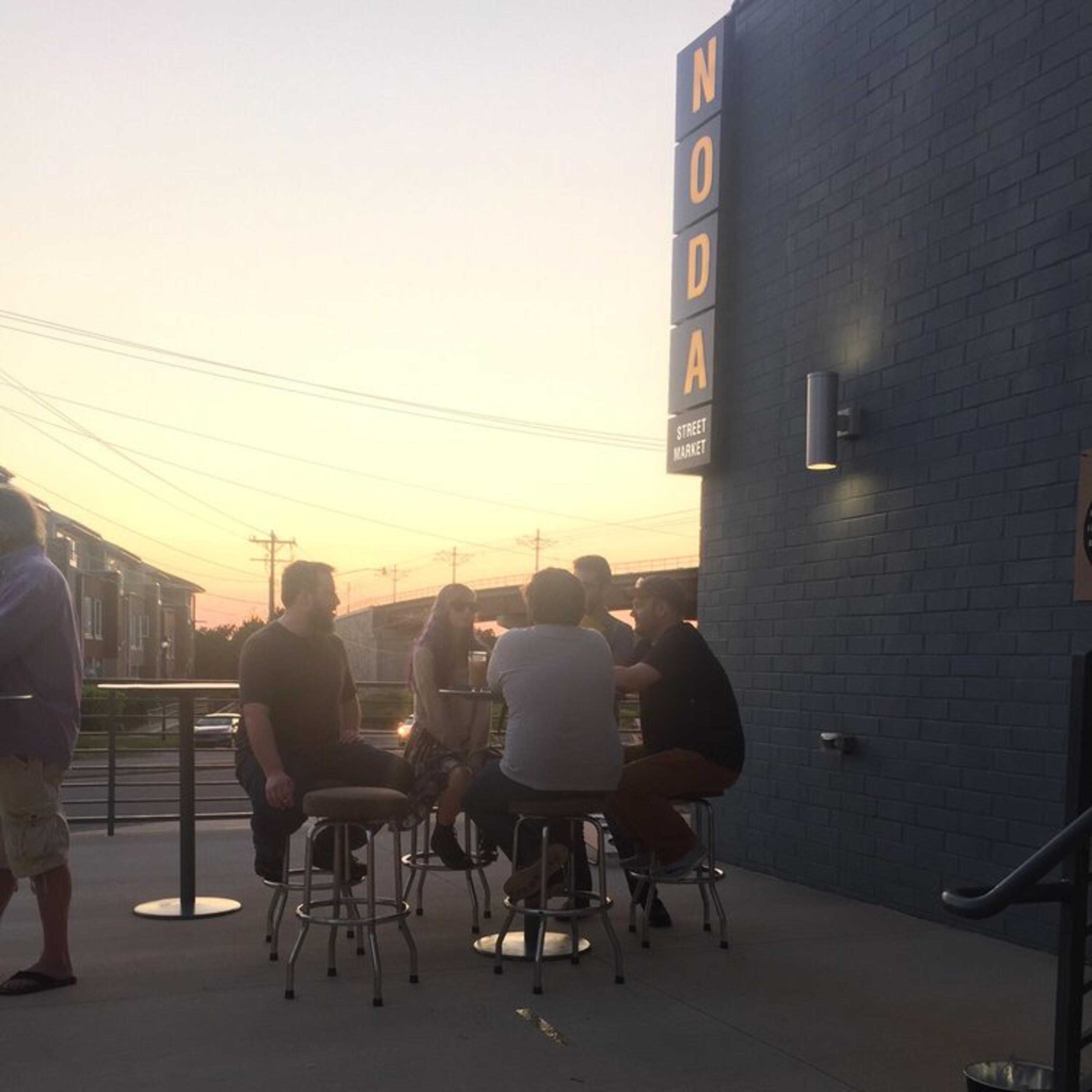 A group of five people sit at high-top tables outside a building during sunset, with power lines and a clear sky in the background.