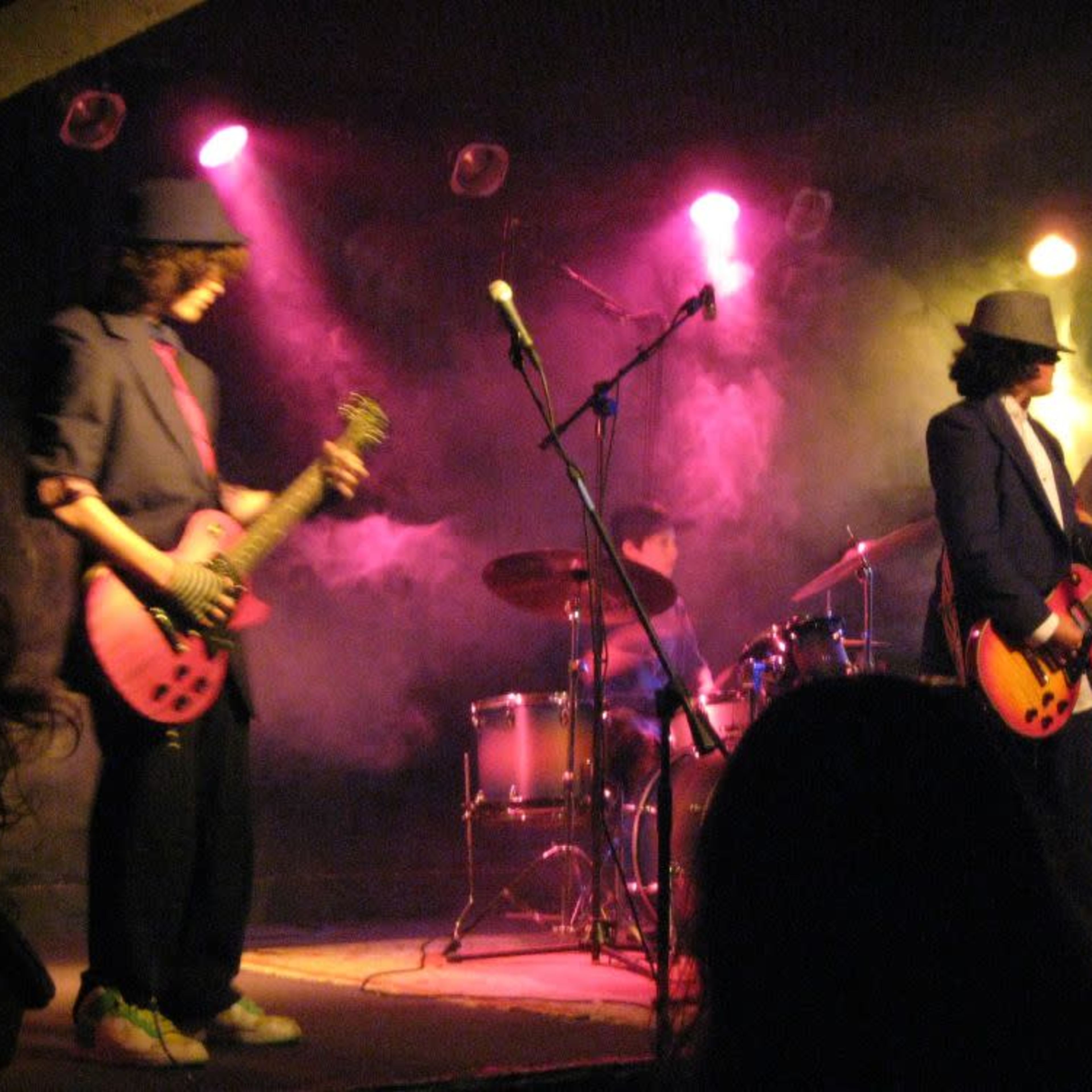 A band performs on stage with electric guitars and a drum set, surrounded by colorful stage lights and fog.