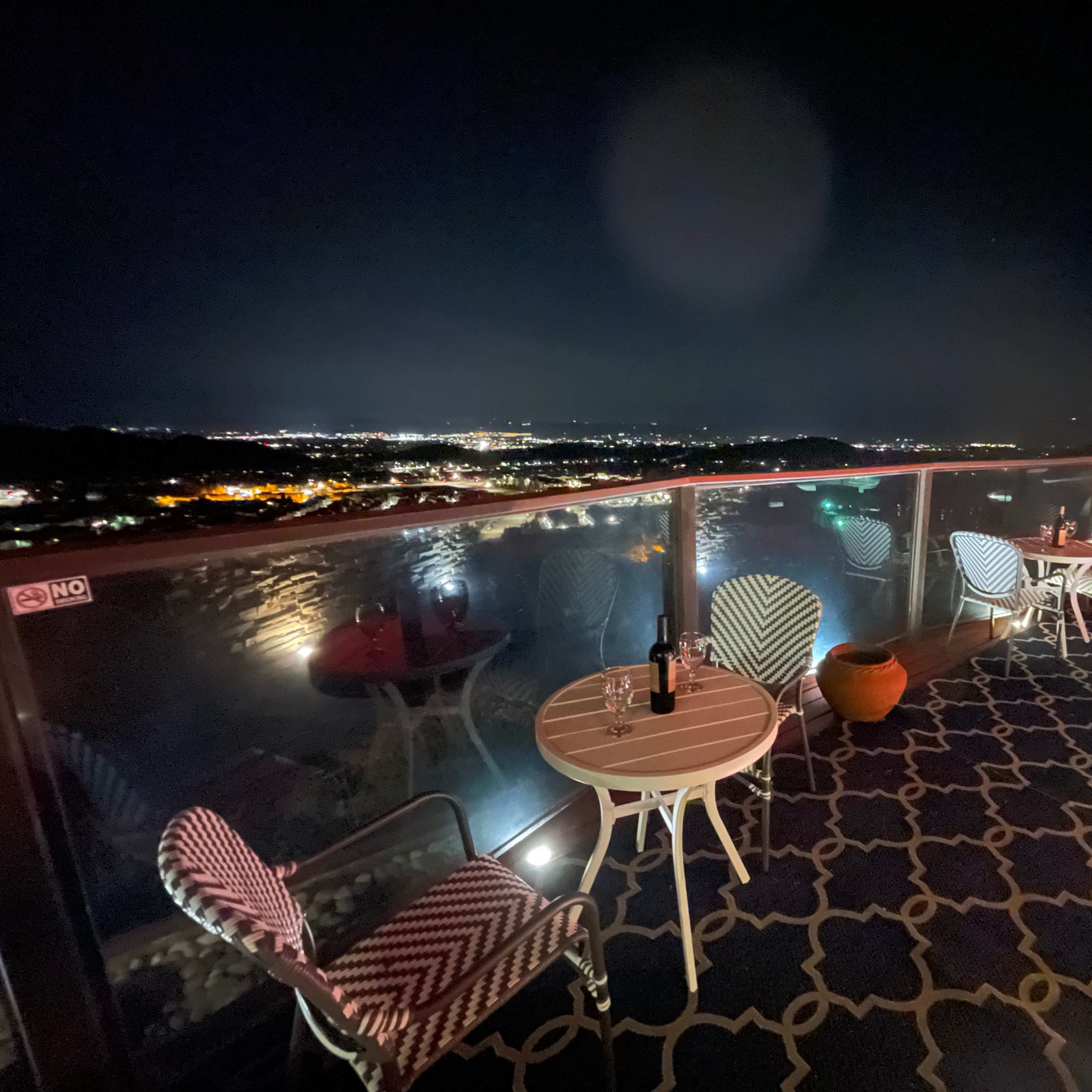 A dimly lit terrace features two chairs and a small table overlooking a city skyline at night.
