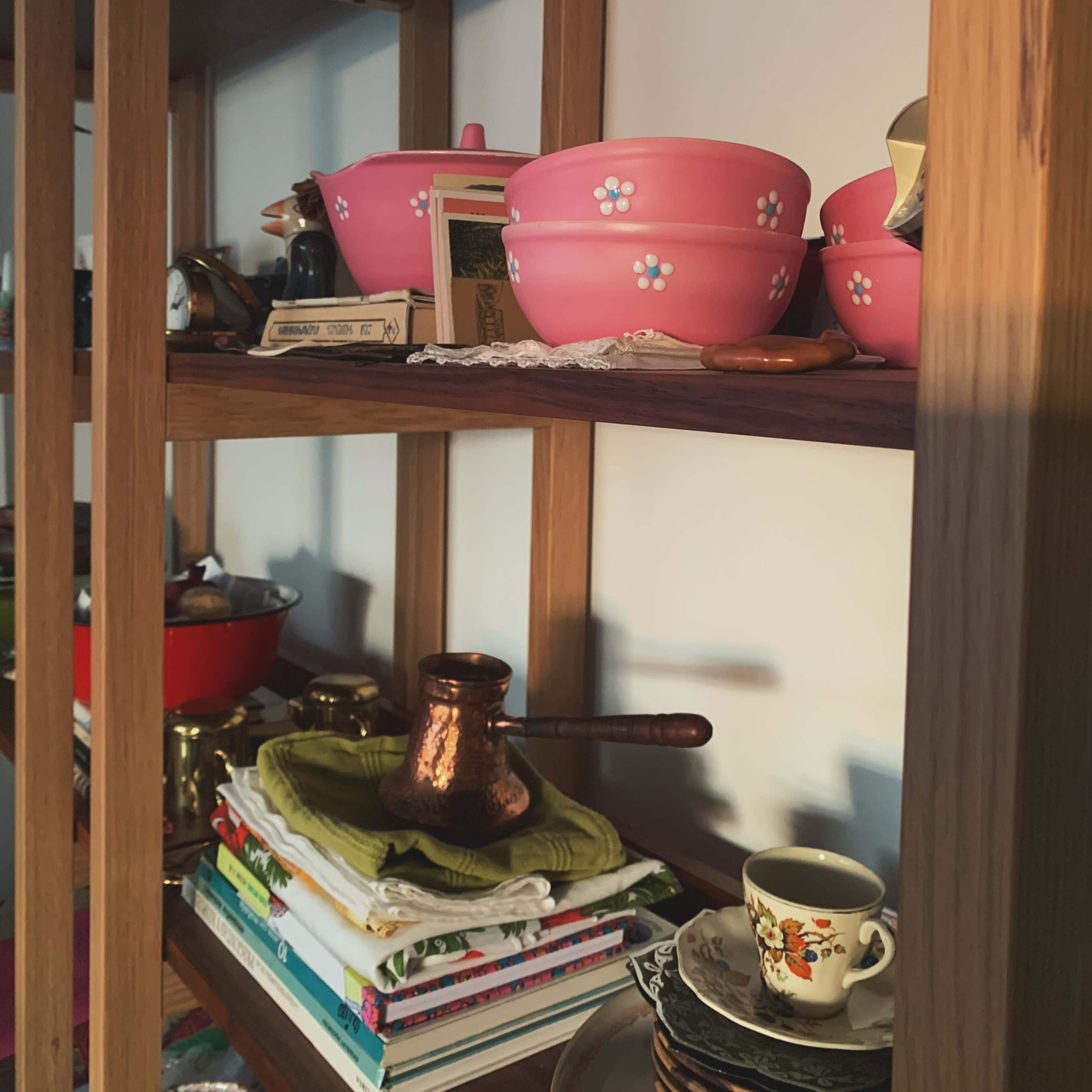 A wooden shelf displays various kitchen items, including pink bowls, a copper cezve, and stacks of magazines.