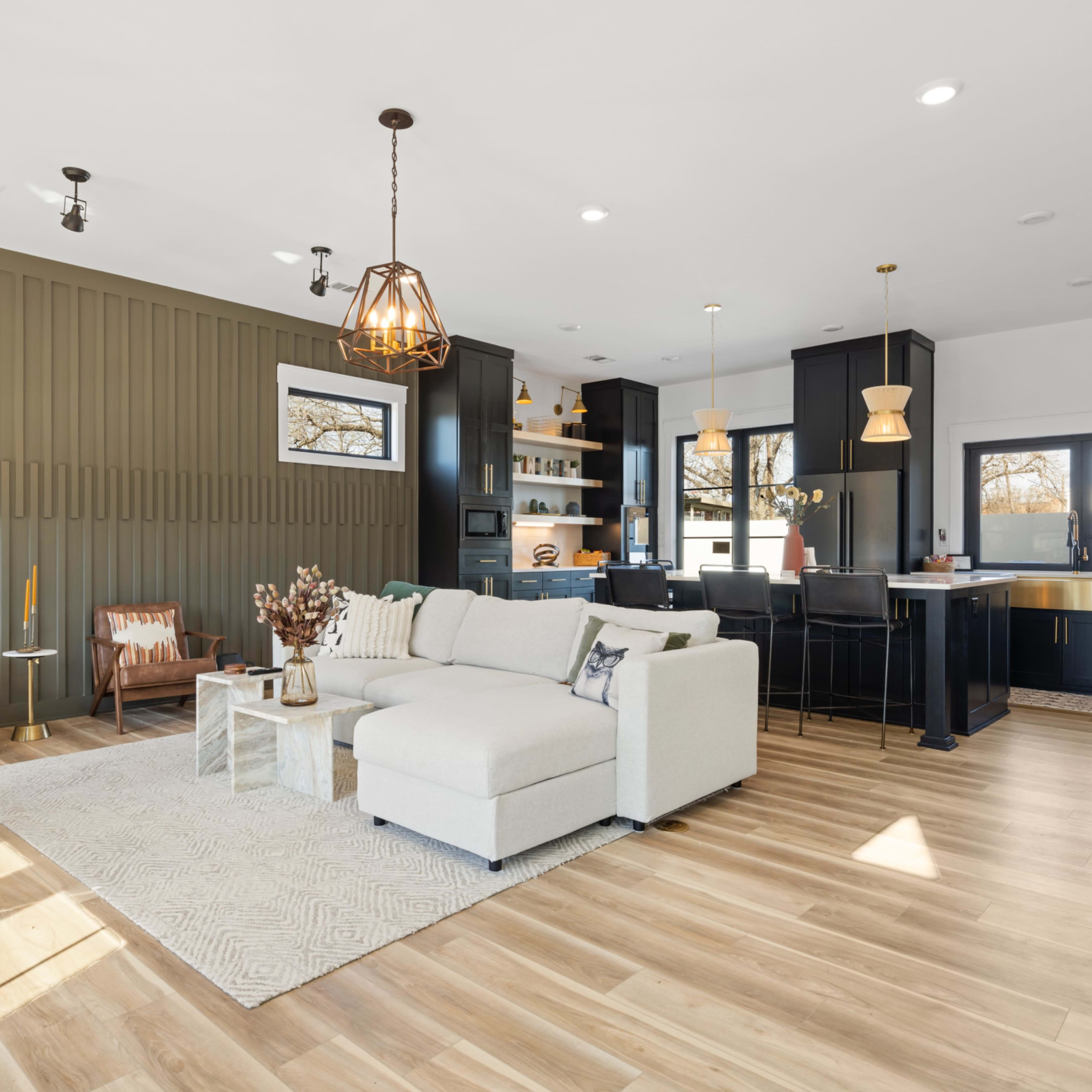 A modern living room features a light sectional sofa, wooden accents, and a dark kitchen area with stylish pendant lighting.