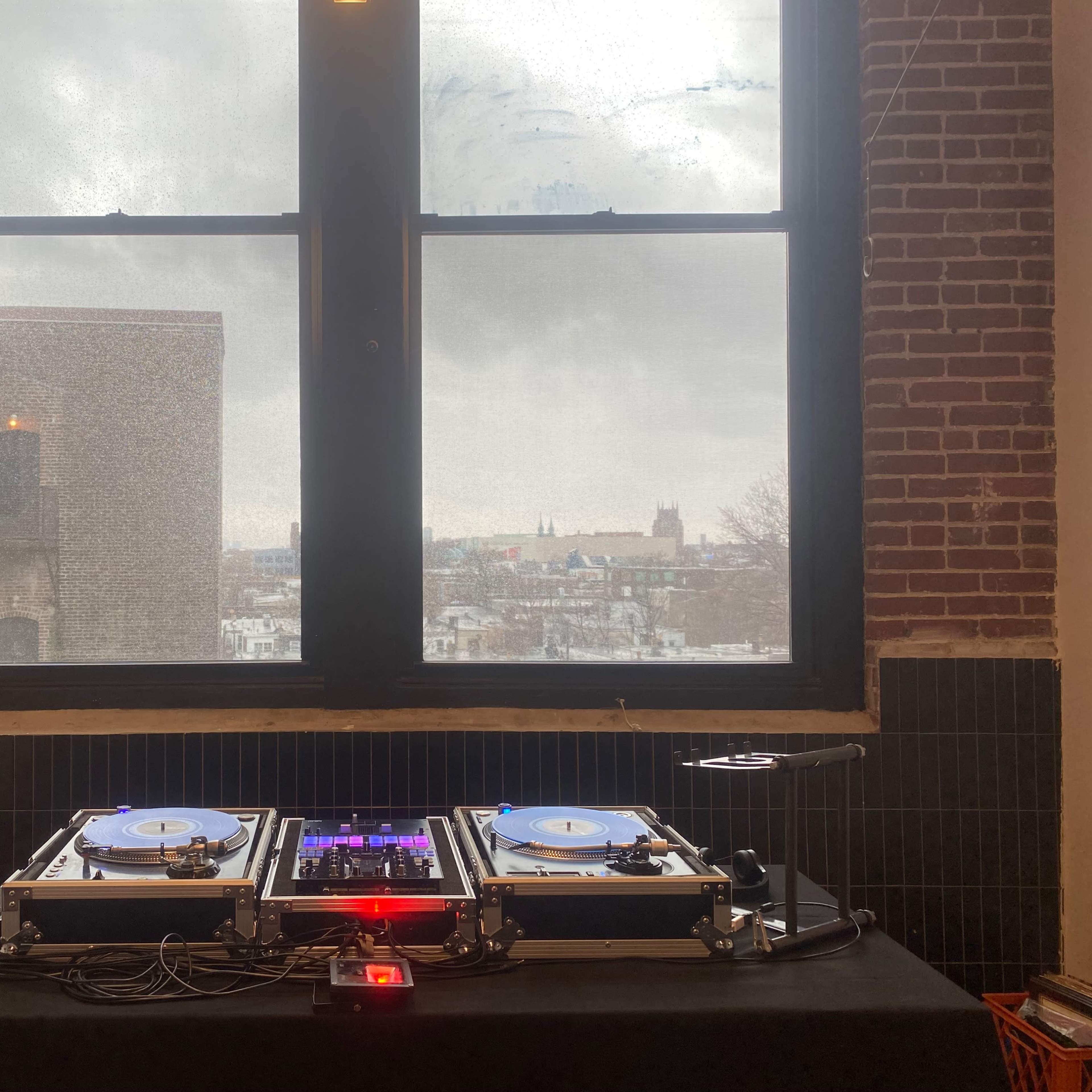 A DJ setup with turntables and a mixer is positioned in front of a large window overlooking a city skyline.